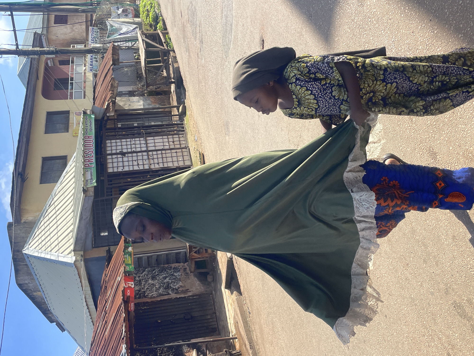 In Jos, Nigeria, on a sunlit street, a woman and a young girl walk side-by-side. The woman is covered in a flowing, olive-green hijab with a white lace trim, and is wearing a blue and orange patterned dress. The girl, to her right, is wearing a dark green headscarf and a yellow, blue, and white floral patterned dress. They are walking on a paved road lined with buildings that appear to be shops or residences, some with corrugated iron roofs and others with more modern construction. Signs with text are visible on some of the buildings, suggesting commercial activity. The scene captures a moment of everyday life on a street in Jos.