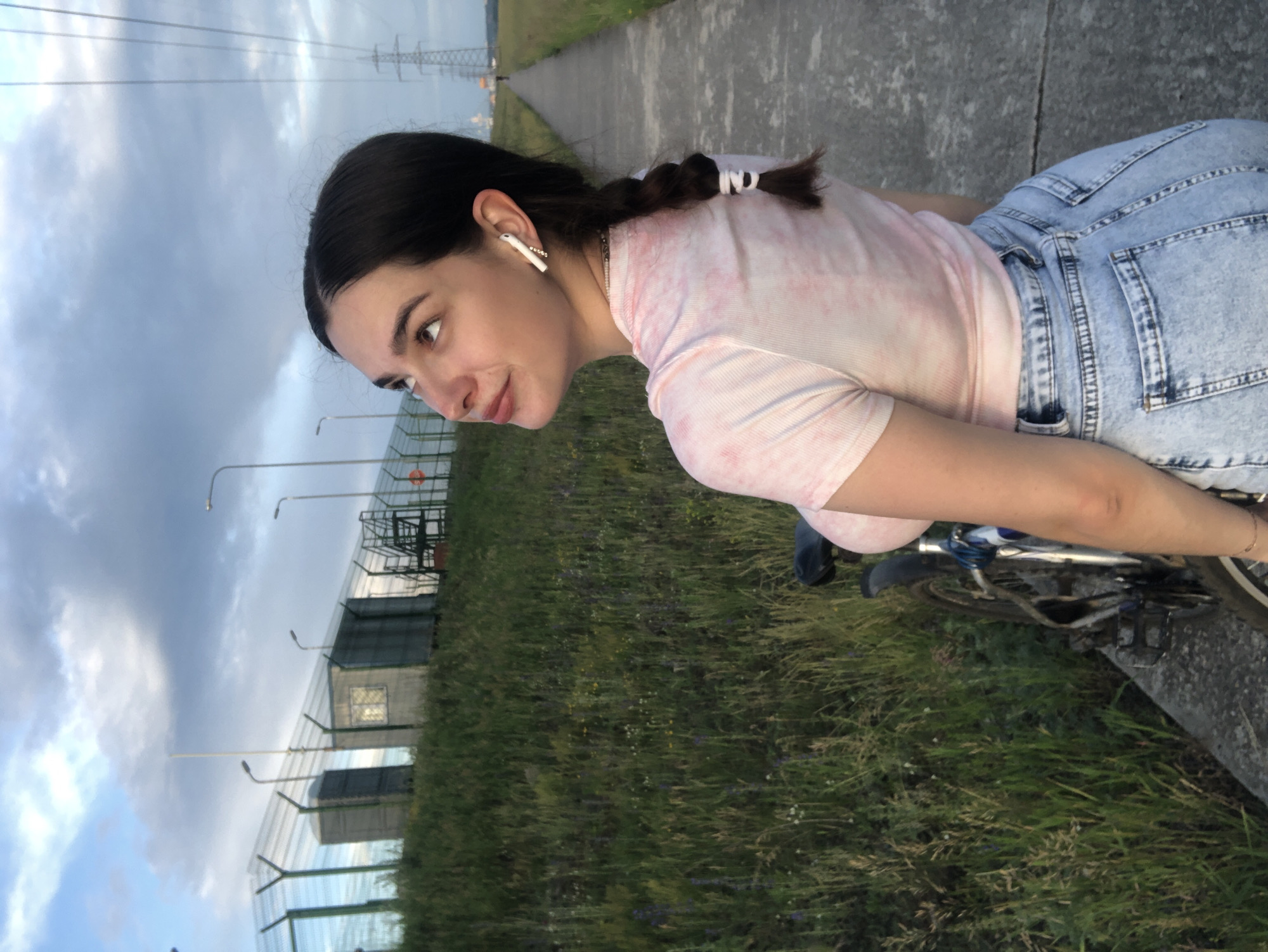 In Noviy, Russia, a young woman with dark hair in a braid, wearing a pink tie-dye t-shirt and light blue denim shorts, stands beside a bicycle. She has white wireless earbuds in her ears and is looking upwards towards a cloudy sky. The scene unfolds on a paved walkway next to a grassy embankment. Behind the embankment, a fence surrounds a complex with several small buildings and industrial structures, possibly a utility area or a facility. In the distance, power lines and transmission towers are visible against the twilight sky. The overall ambiance suggests an outdoor setting in the late afternoon or early evening.