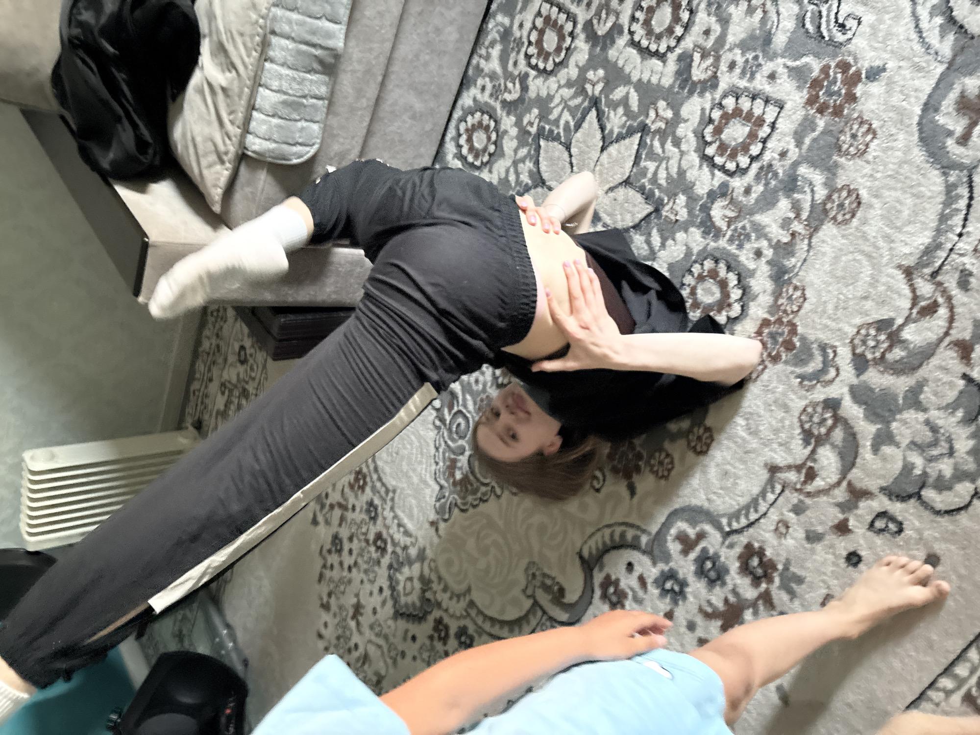 An overhead shot captures a young woman performing an exercise on a patterned rug. She is lying on her back with her legs extended upwards, her torso supported by her arms. The woman is wearing a black t-shirt and black pants with a white stripe down the side, and white socks. Her right leg is bent at the knee, with her foot resting on her upper thigh. The room appears to be a living area, with a gray sofa in the background. A white radiator is visible on the wall, and a black object, possibly a speaker, is to the left of the radiator. The lighting is dim, suggesting an indoor setting. The overall scene is casual and relaxed, with no obvious signs of distress or danger.