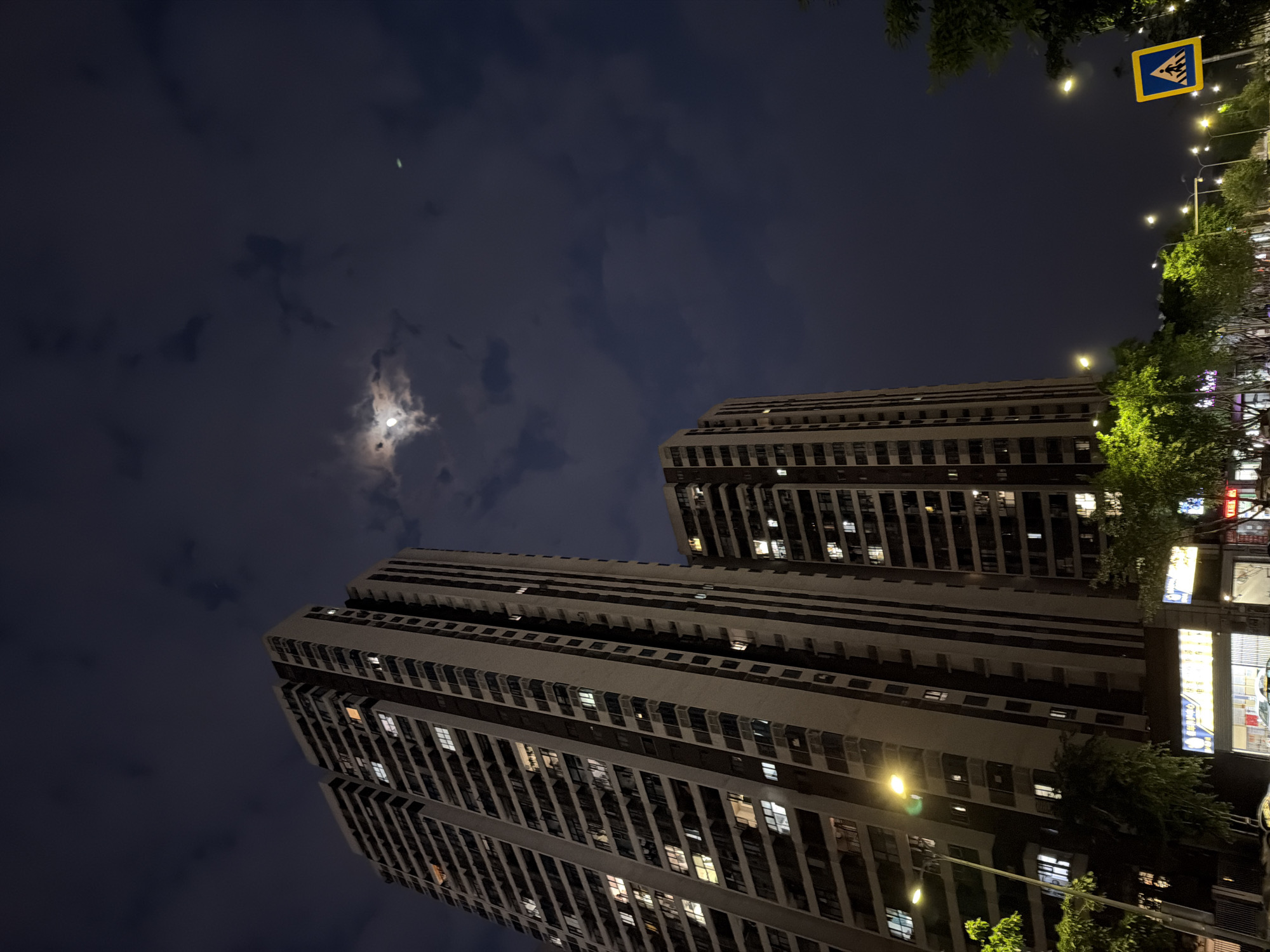 The image, taken at night, shows two tall residential buildings in the Chongqing High-tech Zone, China. The buildings have multiple stories with numerous windows, some illuminated. The sky is dark with clouds and a faint moon visible. Streetlights illuminate the buildings and the surrounding area, including a pedestrian crossing sign at the top right corner of the frame. Trees and some commercial signage are visible on the right side of the image, suggesting a street-level view. There are no people directly visible in the image.