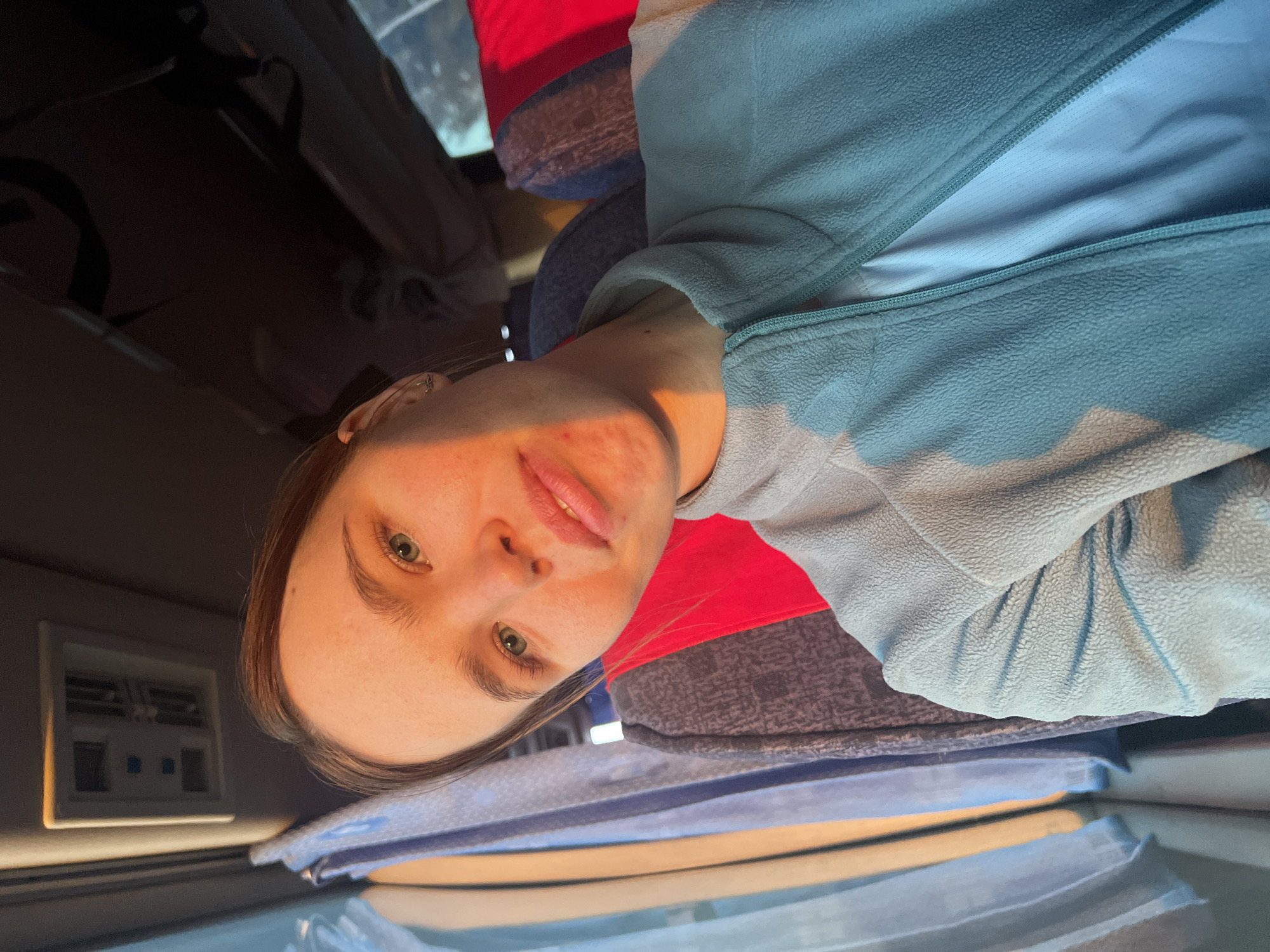 A woman with fair skin, light brown hair, and green eyes is positioned in what appears to be the interior of a vehicle. She is wearing a light blue fleece jacket over a white shirt. The vehicle's interior includes seats with red and grey patterned upholstery, a blue seat covering, and dashboard elements. The exact location in Ustinovo, Russia, cannot be determined from the image alone.