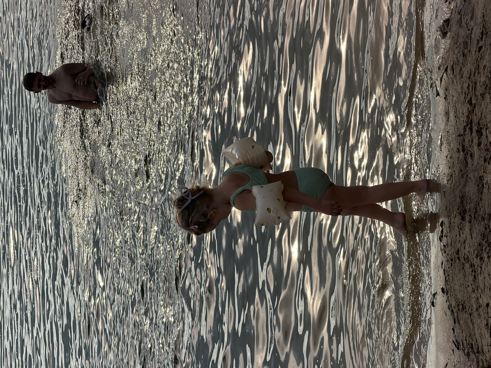 The media file depicts a scene at a body of water, likely a lake or river, in Malakhovka, Russia. The sun is glinting off the water's surface, suggesting it is either early morning or late afternoon. The water is relatively calm with small ripples.
In the foreground, a young girl is standing in the shallow water. She is wearing a light green swimsuit and has a swim float, resembling a small white pillow with an apple design, held in her arms. Her hair is tied back with a light purple accessory. She appears to be looking down at the water or the sandy shore next to her.
In the background, a man is standing in the water, partially submerged. He is shirtless and appears to be facing the camera, with a slight smile.
The overall impression is one of a peaceful, recreational moment at the water's edge, possibly during a family outing. The lighting indicates pleasant weather. There is no visible text in the media file.
