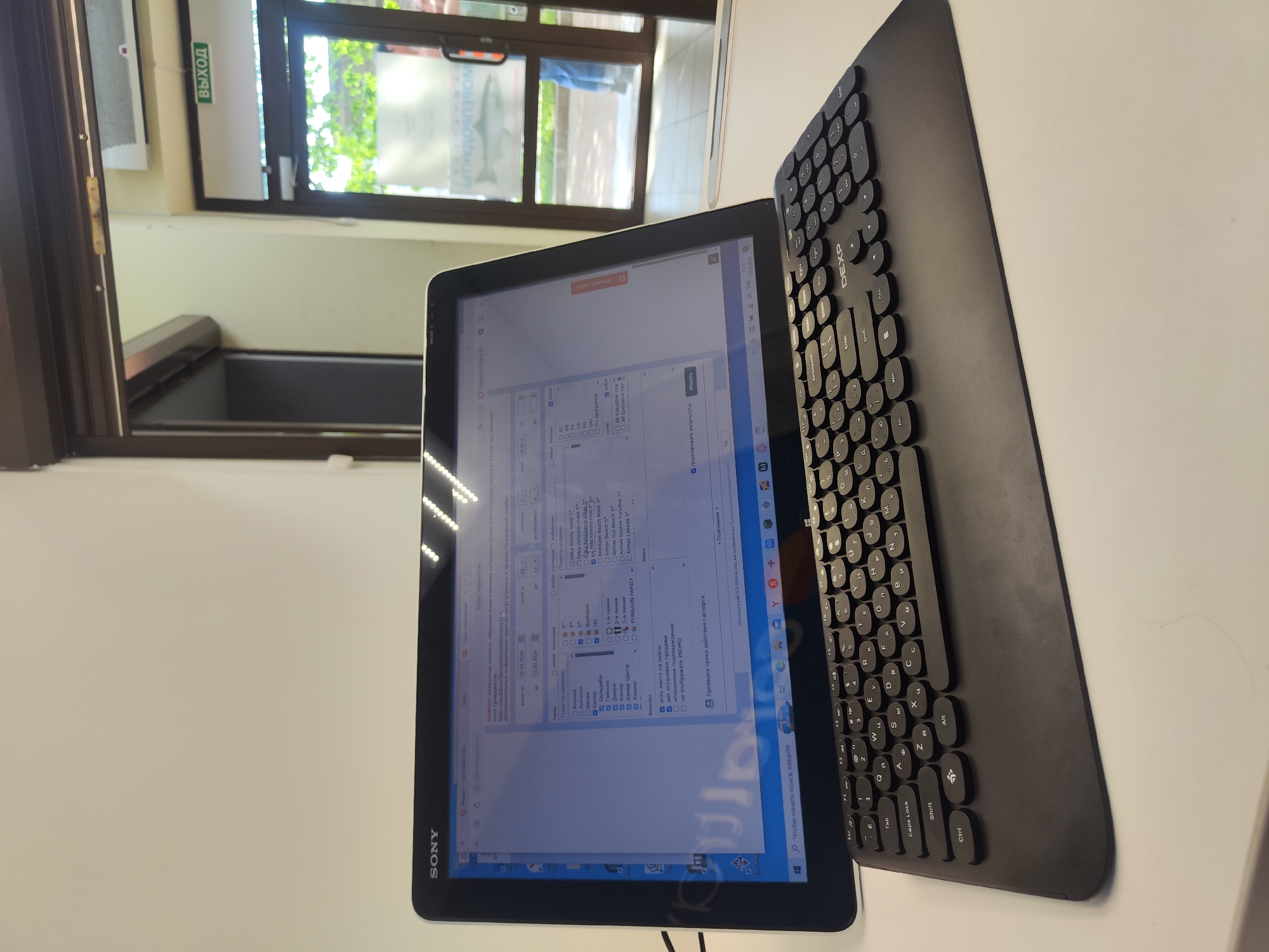 The image shows a Sony tablet computer and a black keyboard on a white surface, likely a table. The tablet displays a search form with text in Russian. A window is visible behind the tablet, showing an exterior scene with greenery. A sign with the word "ВЫХОД" (Exit) in Russian is visible above the window frame. The keyboard appears to be a DEXP brand.
There is insufficient information to determine if the image was taken in Saint Petersburg, Russia.