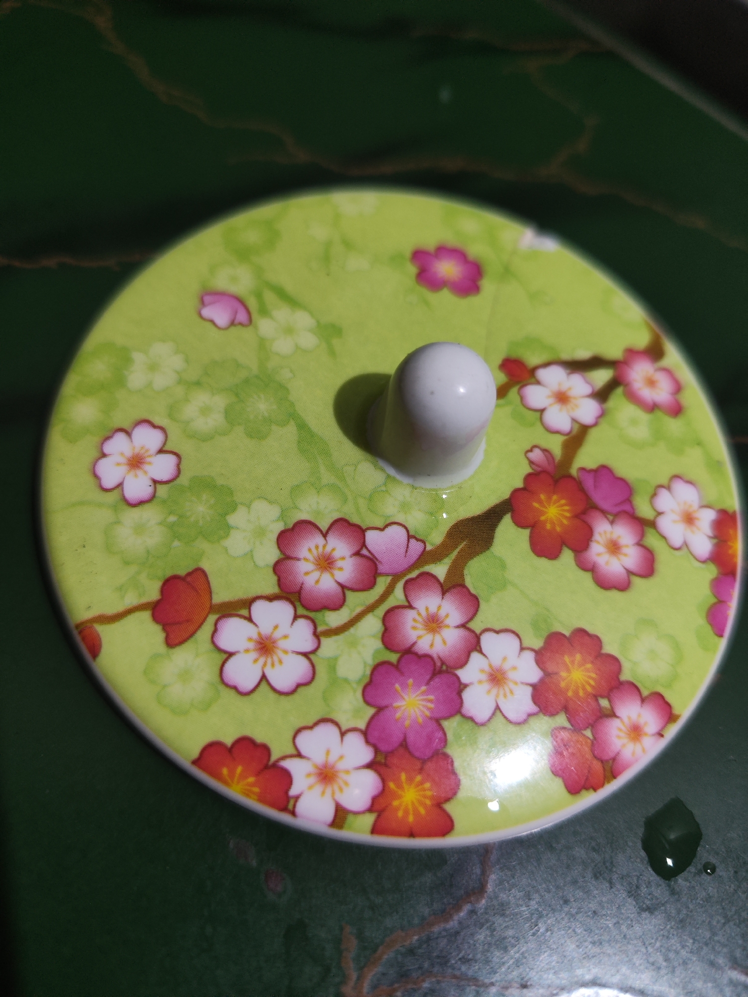 A close-up, overhead shot shows a light green, round lid with a floral pattern featuring cherry blossoms in shades of pink, white, and red, against a dark green, marbled surface. A small, white, rounded knob protrudes from the center of the lid. The lid is slightly angled. The dark green surface has some faint brown marbling and a small, dark, cube-shaped object is visible on the lower right, with a small water droplet next to it. The image's focal point is the lid and its decoration. No people or discernible city elements are present.