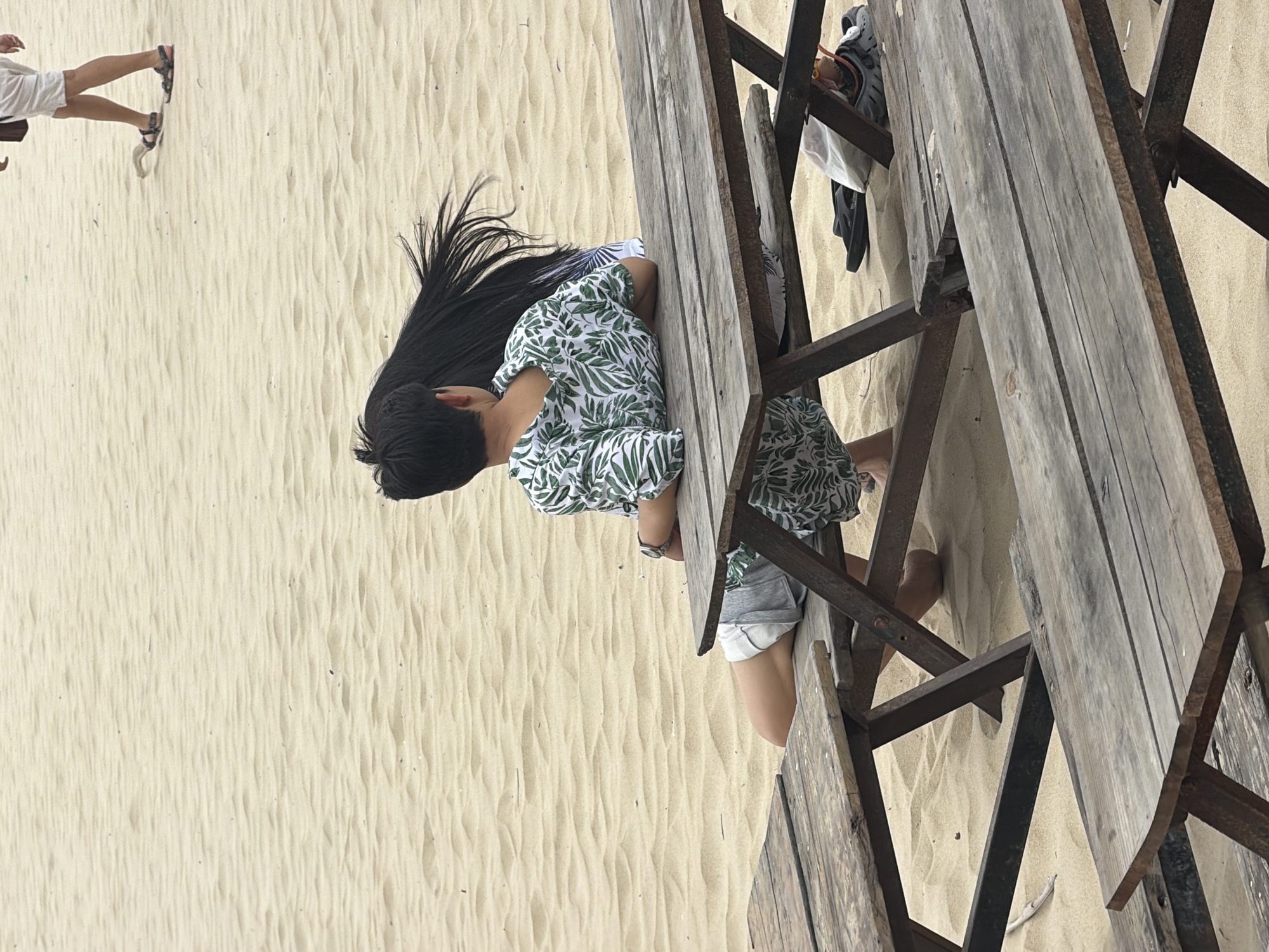 This image depicts a casual outdoor scene set on a sandy beach or dune, likely in Đà Nẵng, Vietnam. The photo is rotated 90 degrees clockwise.
In the foreground, a person with long, straight black hair, wearing a white shirt with a distinct green leaf pattern, is seated at a rustic wooden table. The person's back is to the viewer, and their forearms rest on the table surface, suggesting they are either leaning forward, looking down, or engaged in an activity. A watch is visible on their left wrist. The table itself is constructed from weathered wooden planks supported by dark, industrial-looking metal frames.
Underneath the table, another pair of feet, wearing dark sandals or shoes, are visible on the sand, indicating the presence of at least one other person sitting or resting. In the background, to the left, the lower legs of a third individual are seen, wearing white shorts and dark sandals, standing on the textured sand.
The ground is covered in light beige sand, showing clear ripple patterns across its surface, characteristic of wind or water movement on a beach or dune. The natural lighting suggests it is daytime and likely sunny, contributing to a bright and relaxed atmosphere. The scene evokes a sense of leisure and outdoor dining or resting by the coast. No visible text is present in the image.