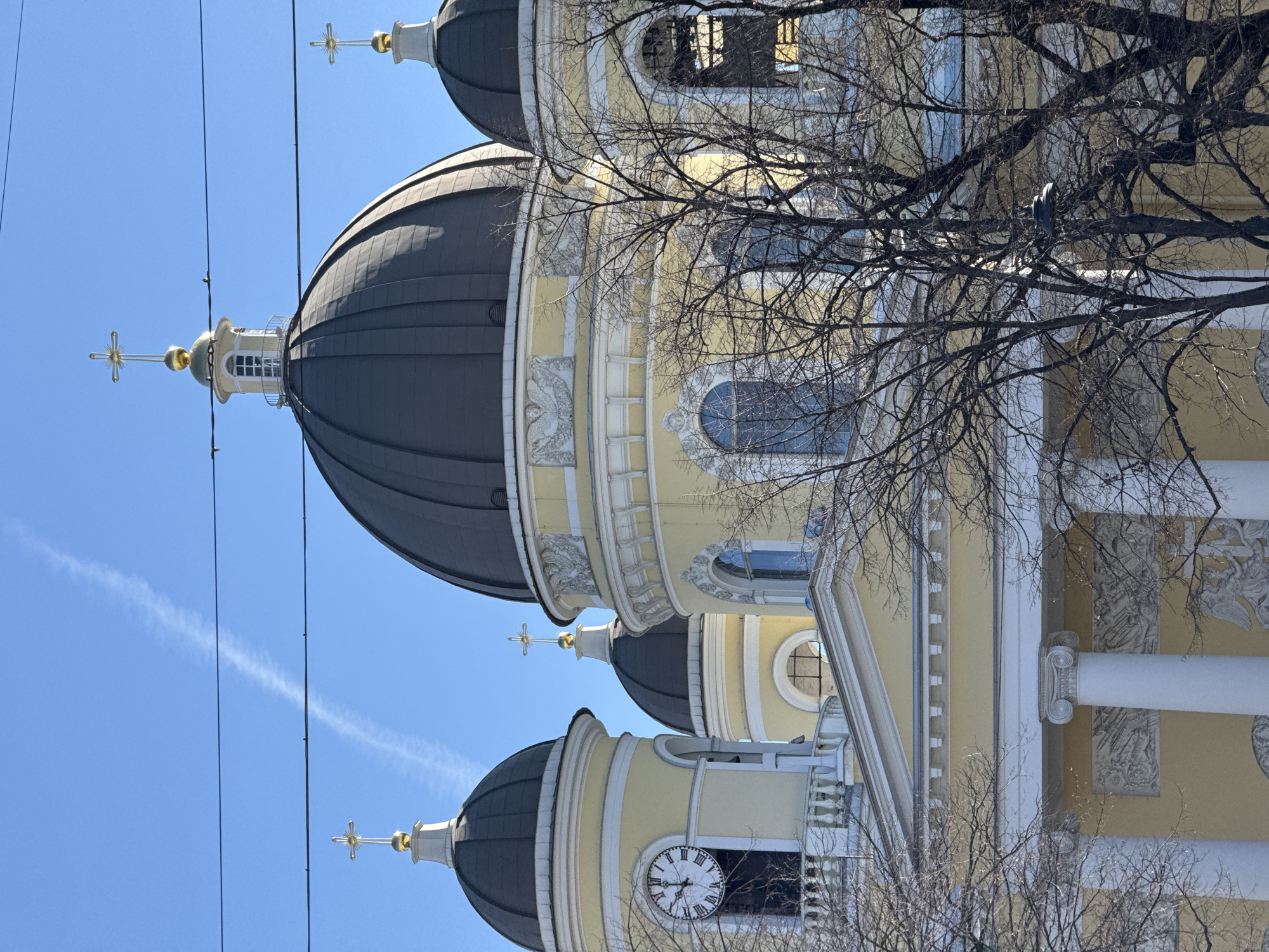 The image shows a section of the Naval Cathedral of Saint Nicholas in Saint Petersburg, Russia. It depicts the domes, architecture, and clock of the cathedral against a clear blue sky, partially obscured by bare tree branches. Visible architectural elements include ornate columns, arched windows, and religious crosses atop the domes. A clock tower with a white clock face is seen closer to the bottom of the image. Electrical cables are visible in the left of the frame.