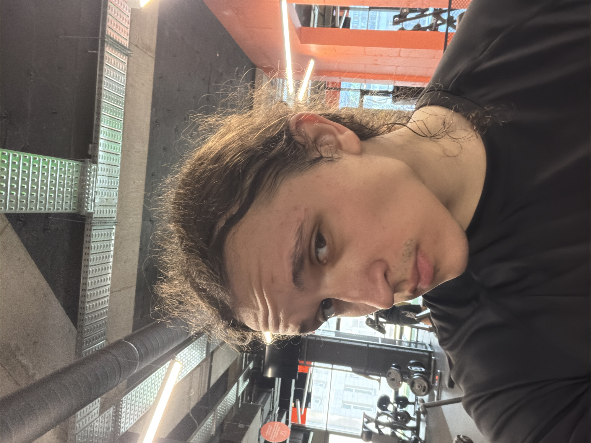 A person with long, wavy brown hair and light skin is oriented in a gym environment. The individual is wearing a black t-shirt. The gym has a variety of equipment, including weight machines. The ceiling features orange support beams and overhead lighting. Ventilation pipes and grated fixtures are visible on the left side. Through the windows, buildings and the sky are seen in the background. I cannot verify the location is in Plovdiv, Bulgaria.