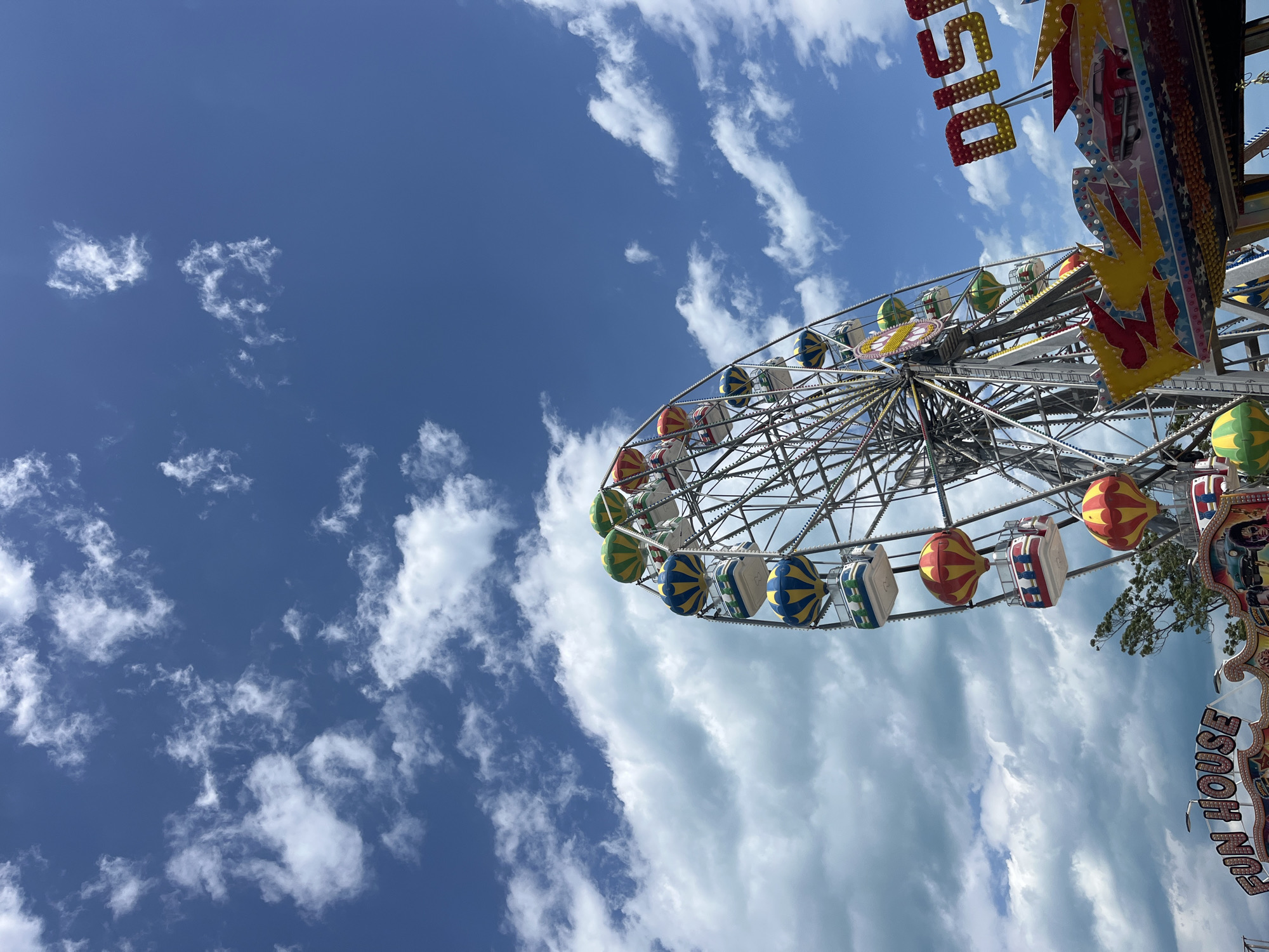 The image shows a Ferris wheel at a fair or amusement park under a blue sky with scattered clouds. The Ferris wheel has enclosed gondolas in various colors (green, blue, red, and white). The "DISCO" sign is visible above the Ferris wheel. A "FUN HOUSE" sign can also be seen. The location is likely in Varna, Bulgaria, as the city has several amusement parks and fairgrounds along the coast.