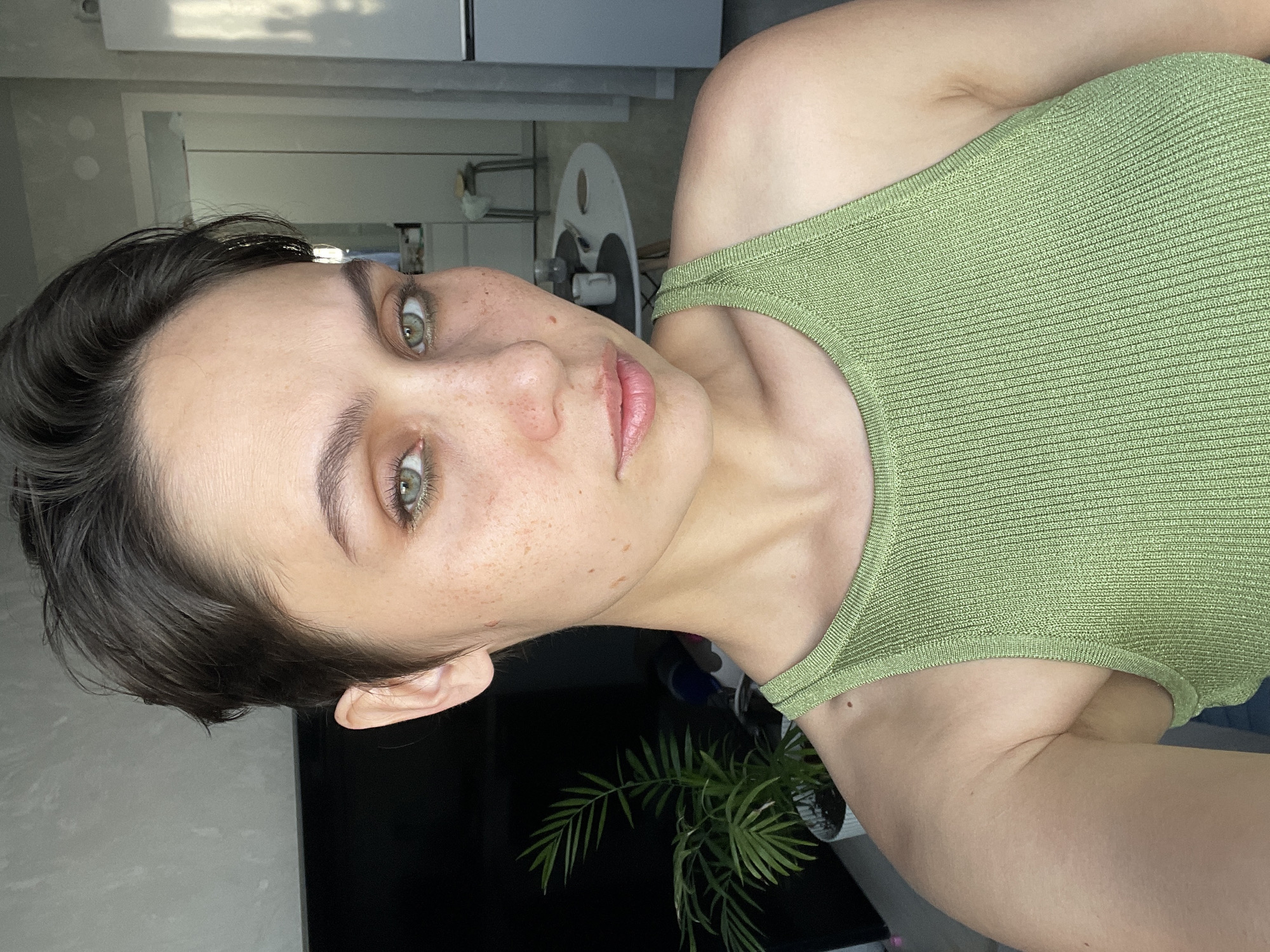 A woman with short dark hair and green eyes is taking a selfie. She is wearing a light green knit top. Her skin is fair with freckles. The background is a blurred interior of a room, partially showing kitchen cabinets, a table, and a potted plant. The location is not identifiable as Izhevsk, Russia from the image alone.