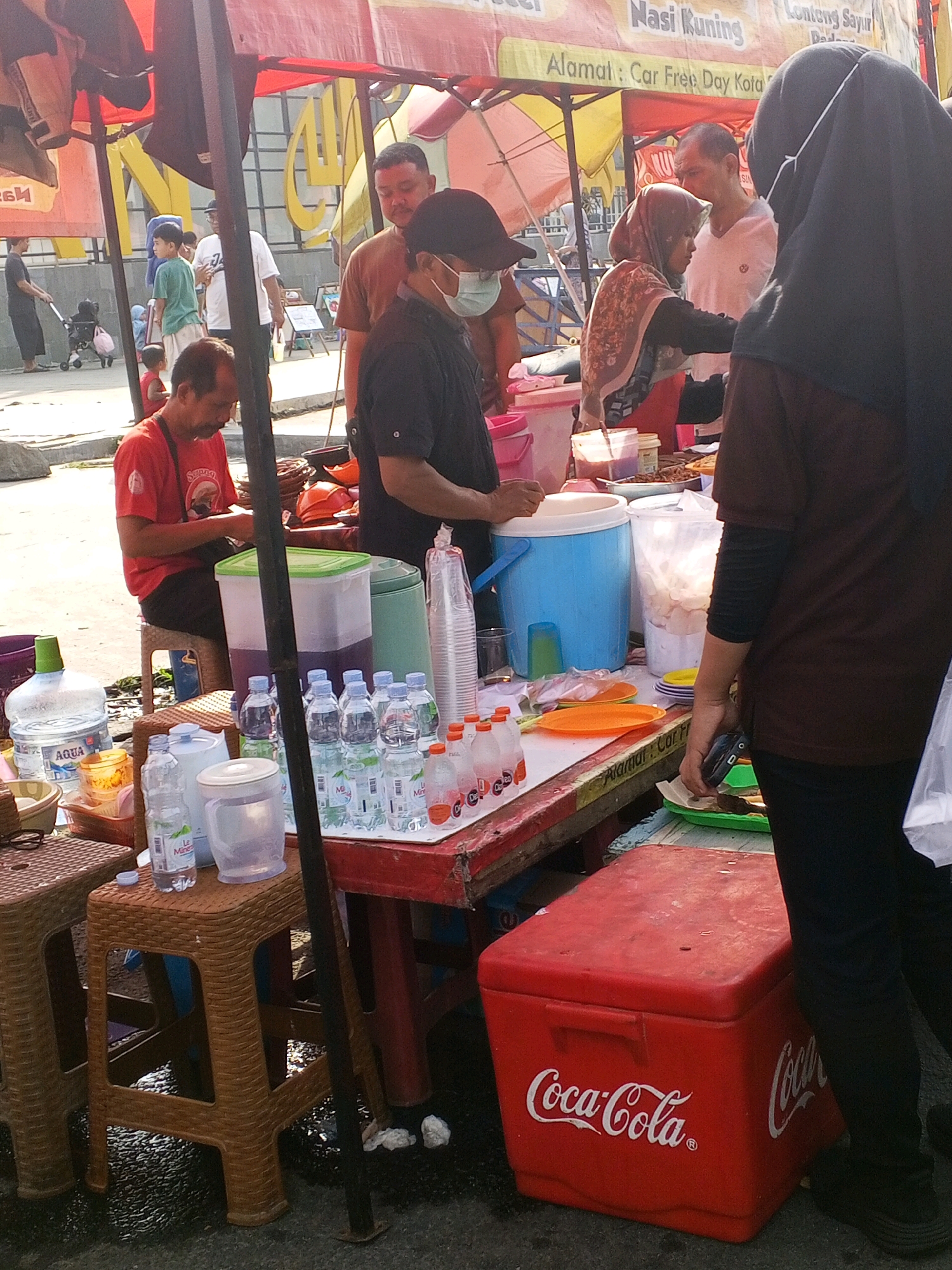 A busy outdoor market stall is depicted in Serang, Indonesia, with a variety of food and drink items on display. In the foreground, a red Coca-Cola cooler is prominent. To its left, several plastic bottles of water and other beverages are arranged on a table. A man in a red shirt is seated behind the table, appearing to be serving customers. Further back, a woman in a headscarf is interacting with a customer. The stall is covered by a colorful awning, and the ground is wet, suggesting recent rain. The background shows more stalls and a crowd of people, indicating a lively market atmosphere.