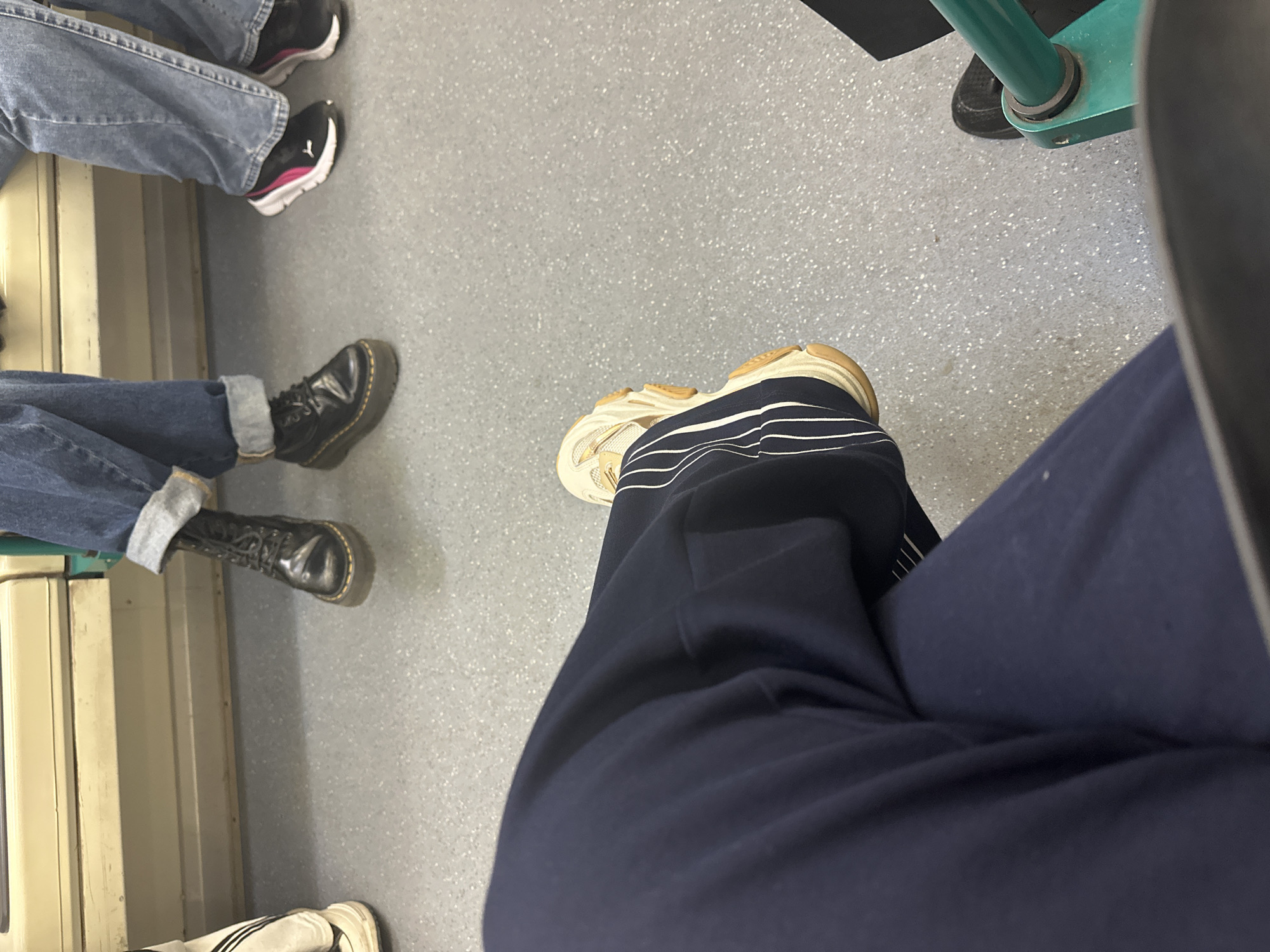The image depicts the interior of a public transportation vehicle. Two individuals are visible from the knees down. One is wearing a pair of Dr. Martens boots and rolled up jeans. Another is wearing denim jeans and Puma sneakers. A third person wearing blue sweatpants and white sneakers is sitting down on a seat, with a pole next to the seat. Based on the interior of the vehicle, it is likely a tram or bus within the city of Sofia, Bulgaria. The floor is speckled gray.