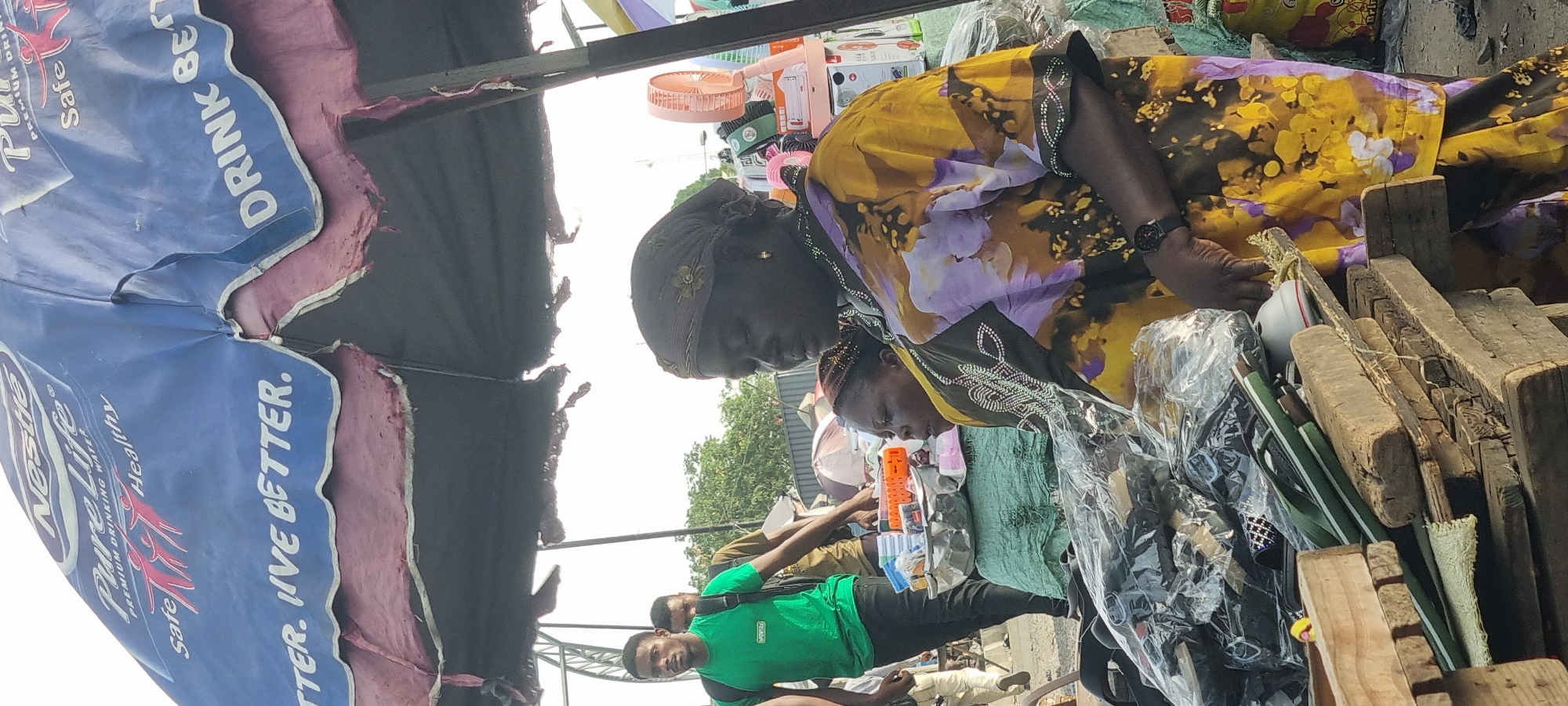 This outdoor image shows a market scene in what appears to be Lagos, Nigeria. Two African women, one wearing a yellow and purple floral top, are focused on items displayed on a wooden surface. One has on a head covering. A man wearing a green shirt is visible in the background. Items for sale include white containers and long green objects. Overhanging the scene is a partially damaged blue umbrella with marketing for "Nestle Pure Life" drinking water. The overall setting suggests a bustling market environment.