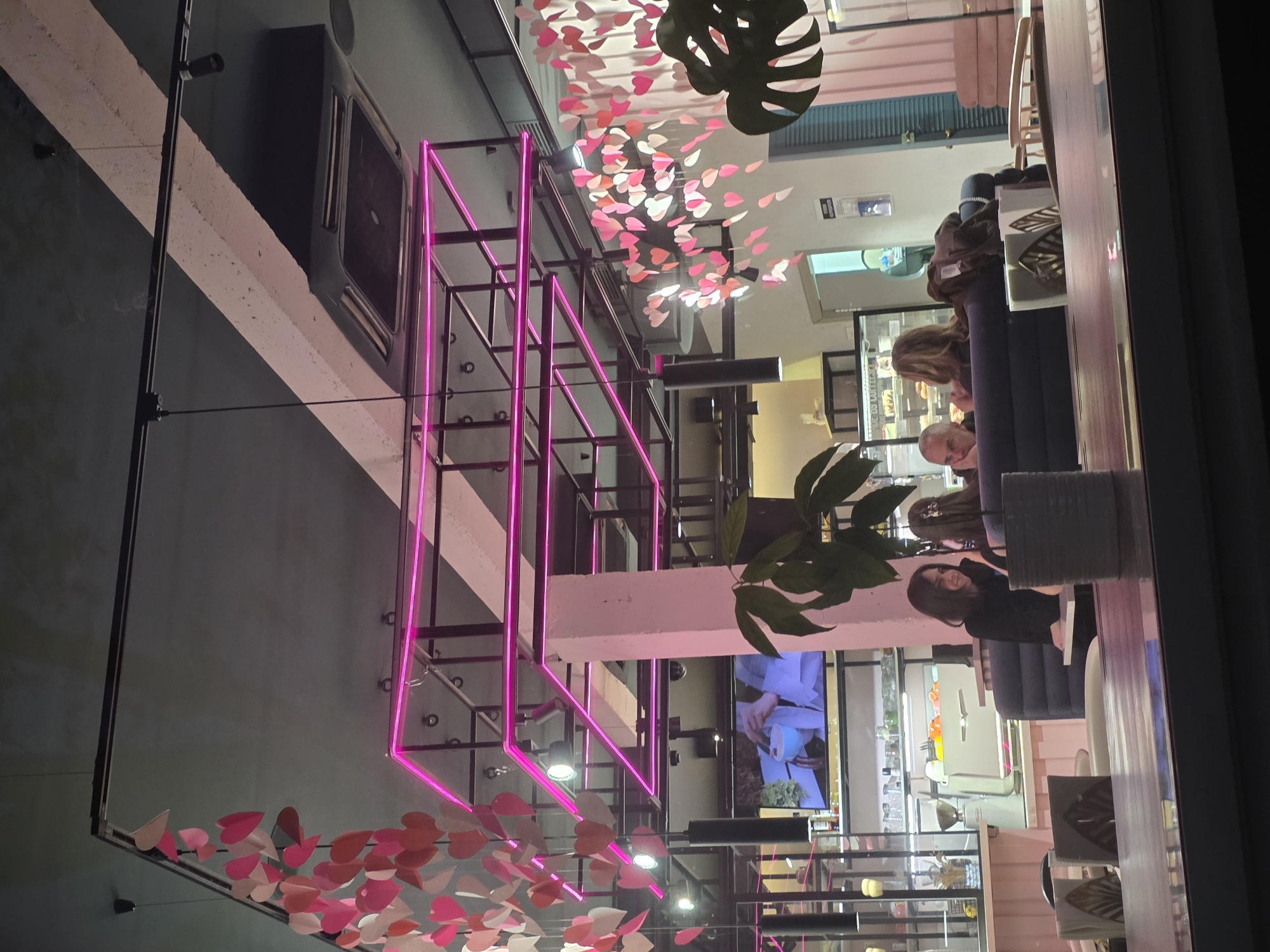 The image shows the interior of a cafe or restaurant in Krasnodar, Russia, as viewed through a window. The scene is characterized by modern decor featuring a pink neon geometric structure, a ceiling decoration of paper hearts, and potted plants. Patrons are seated at tables, including at least four people visible, one of whom is looking towards the camera. Various objects such as chairs, tables, glassware, refrigerators, and a television screen are also visible within the cafe. The overall atmosphere suggests a contemporary and stylish environment.