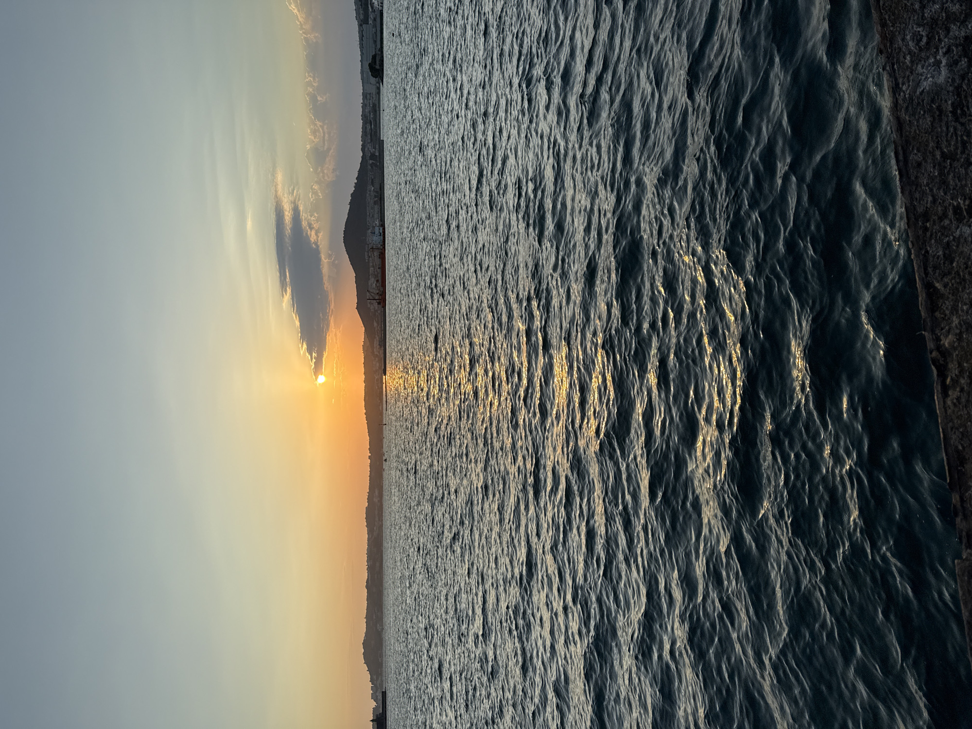 The image depicts a sunset over the sea in Pozzuoli, Italy. The sun, partially obscured by a cloud, casts a warm golden glow across the sky and reflects on the rippling water. In the distance, the coastline of Pozzuoli is visible, featuring a gentle hill topped with buildings and some industrial structures, including a red crane. The foreground is dominated by the dark, textured surface of what appears to be a stone embankment or pier. The overall scene conveys a tranquil and picturesque coastal atmosphere at dusk.