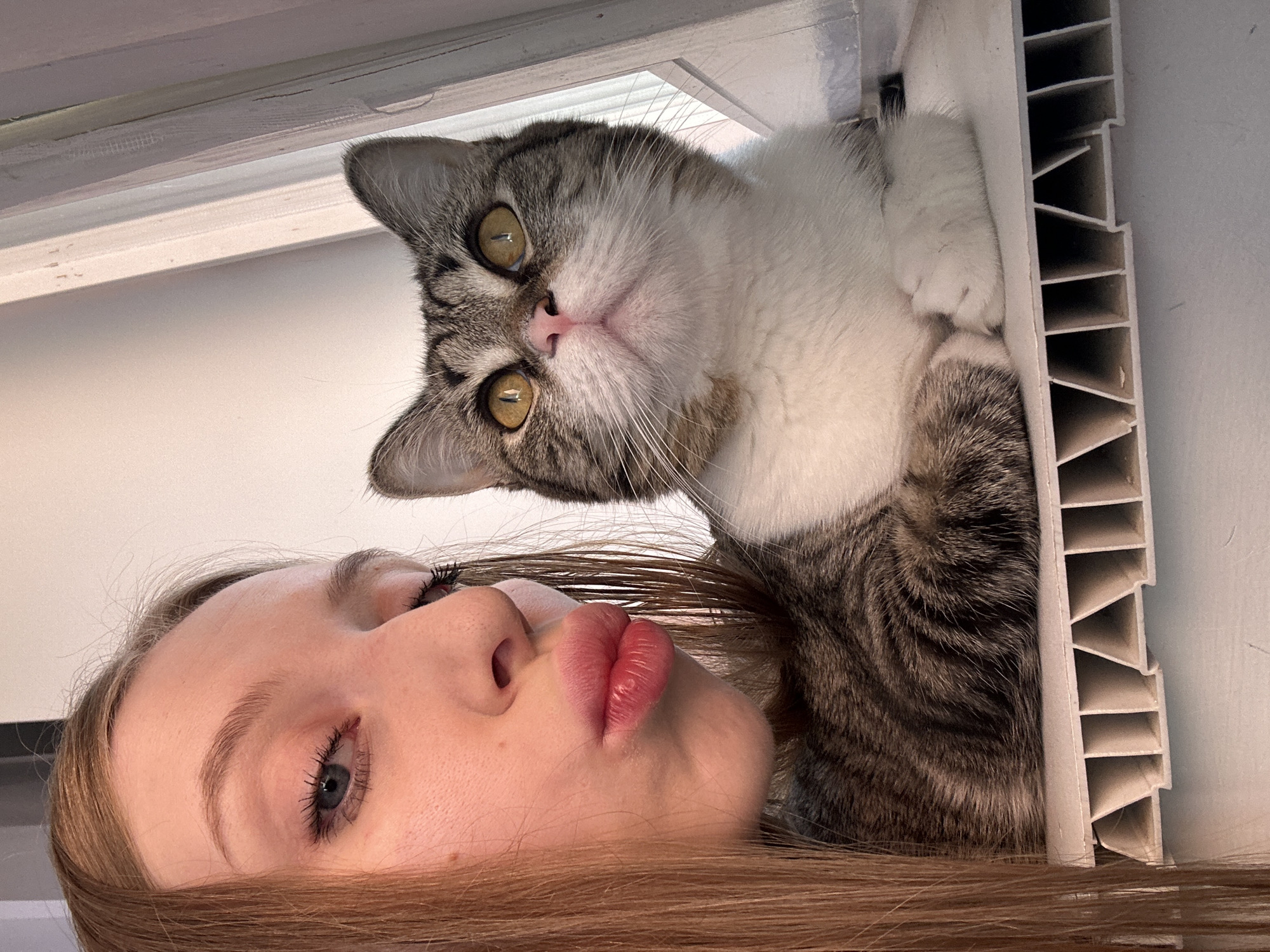 A woman with long, light brown hair is lying on her back next to a gray tabby and white cat. The cat is lying on a white, slatted object, possibly a window air conditioning unit or a radiator cover. The woman is looking up with her eyes partially closed and her lips pursed. The cat is looking down at the camera with its golden eyes. The background is a white wall or ceiling. The location is Chaikovsky, Russia.