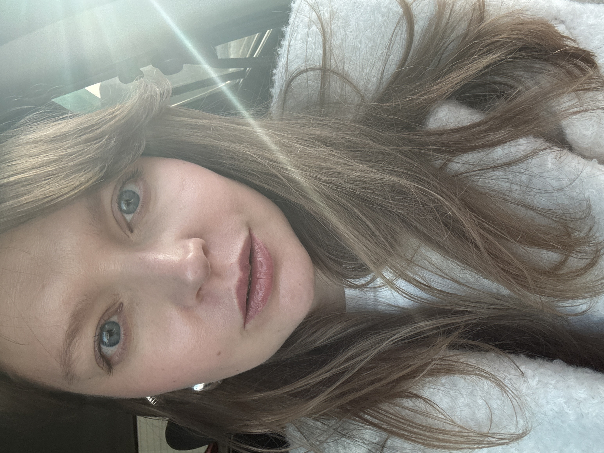 An adult female is depicted in a close-up, chest-up portrait. She has light skin, long, light brown hair parted to her left and falling over her shoulders, blue eyes, a straight nose, and light pink lips that are slightly parted. A silver-colored, crescent-shaped earring is visible on her left earlobe. She is wearing a white, textured garment, appearing to be a fleece or boucle sweater or jacket. Her gaze is directed straight at the camera. The background indicates she is inside a vehicle, with dark structural elements visible on the right and a darker car seat or headrest behind her left shoulder. Strong natural light, consistent with direct sunlight, enters from the upper right, causing significant lens flare. Reflections are visible in both irises. The location of the scene is Izhevsk, Russia.
