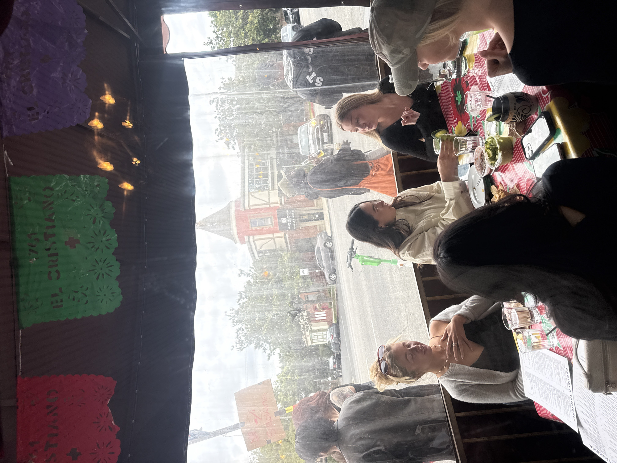 The image shows a group of people sitting at a table, seemingly at a restaurant, with a view of the street outside. There are several people, their faces and bodies partially visible, engaged in conversation and enjoying food and drinks on the table. The table setting includes glassware, dishes, and a menu. Through the window, one can see a street scene with buildings and vehicles, a typical urban setting. Based on the "Escuela Taqueria" menu, the location is Los Angeles, United States of America.