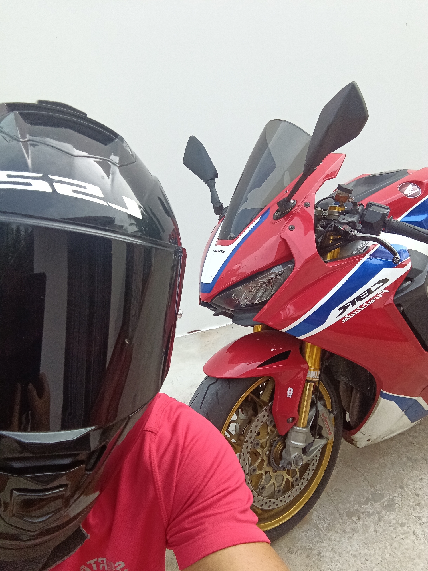 The image displays a person wearing a black full-face helmet and a red short-sleeved shirt, positioned next to a red, white, and blue sport motorcycle. The setting is outdoors in Mũi Né, Vietnam, against a plain, light-colored wall and a concrete ground, under bright daylight conditions.
The person is shown from the chest up, with their face fully obscured by the helmet's dark visor. A white, stylized logo, readable as "S52" from this angle, is visible on the side of the helmet. Their red shirt has a distinct texture.
The motorcycle, identified as a Honda CBR series sportbike, is prominently featured on the right side of the frame. It sports vibrant red fairings accented with white and blue racing stripes. Notable details on the bike include a clear headlight, a dark-tinted windscreen, and black rearview mirrors. The handlebars show a small brake fluid reservoir. The front wheel is equipped with a gold multi-spoke rim, gold inverted Ohlins front forks, and Brembo brake calipers. Visible text includes the "Honda" wing logo on the fairing and stylized "CBR" lettering. The motorcycle appears stationary, suggesting the person is posing with it for a photograph.