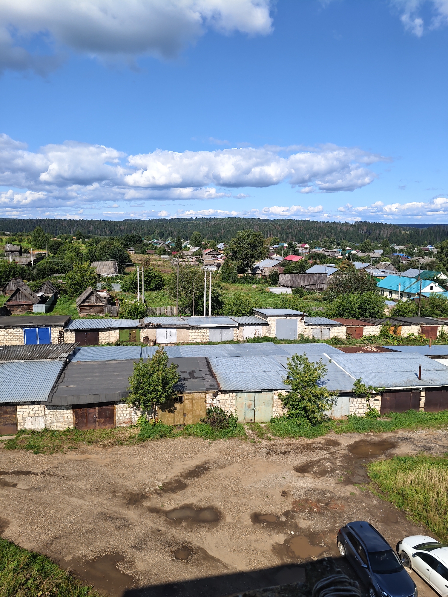 The image depicts a wide view of a rural settlement on a sunny day. In the foreground, there are several rows of garages constructed with brick bases and corrugated metal roofs. The garages are of varying ages and conditions, with some showing signs of rust and weathering. In front of the garages, there is a dirt and gravel area with puddles from recent rain. Two cars, a dark SUV and a white sedan, are parked in this area.
In the midground, the scene transitions to a village with scattered houses of various architectural styles, surrounded by lush green trees and vegetation. Some houses have colorful roofs, suggesting a typical rural Russian landscape. Further in the background, a dense forest stretches across the horizon, meeting a bright blue sky filled with white cumulus clouds. The overall impression is one of a peaceful, sunny day in a rural Russian setting, likely the village of Kurashim.