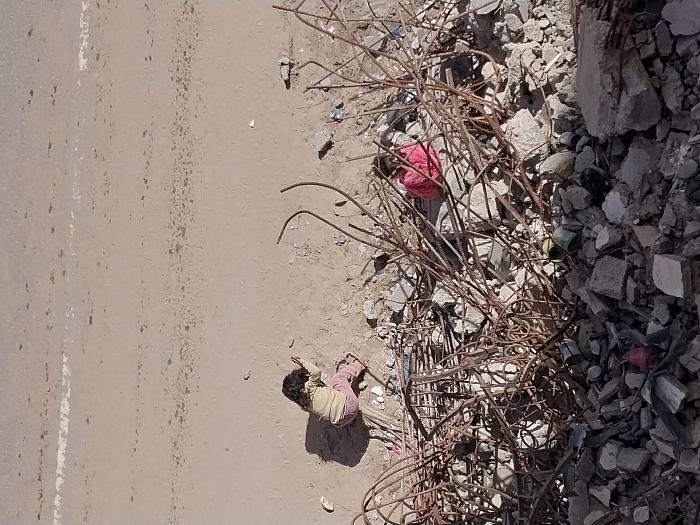 The image depicts two young children amidst a scene of destruction and sandy ground in Khan Yunis, Palestinian Territory. In the foreground, a substantial pile of gray concrete rubble and tangled, rusted rebar dominates, with scattered debris including cans and fragments. Partially obscured by this rebar, a child wearing a bright pink garment is seated or crouching. Further back, on a light brown, unpaved sandy surface, another child, with dark hair, wearing a light-colored top and pink trousers, is positioned prone, appearing to interact with the ground. The background consists of a wide expanse of the same unpaved sandy ground, featuring faint, irregular white markings. The scene is illuminated by overhead sunlight, casting shadows.