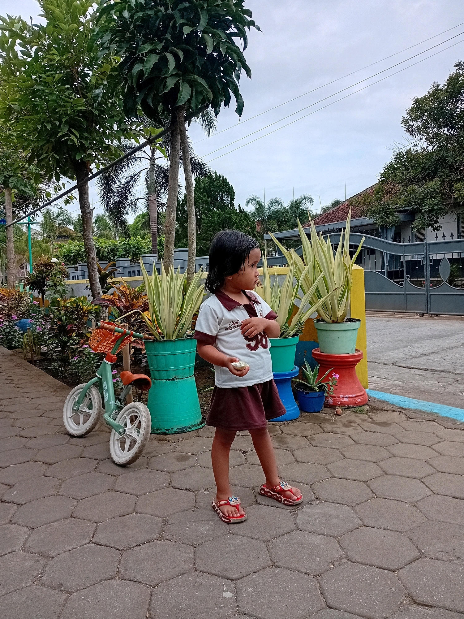 A young girl stands on a paved walkway, holding a small, light-colored object in her right hand. She is wearing a white shirt with red trim and the number "96" on it, and a maroon skirt. Her left foot is adorned with a red flip-flop featuring a cartoon character. To her left, a small, mint-green bicycle with orange handlebars and a brown seat is parked on the walkway. Behind the girl, several potted plants are arranged on colorful stands: two large, light-green, spiky plants in teal pots, one on a blue stand and another on a red stand, with a smaller plant in a blue pot at the base of the red stand. Further back, a variety of other plants and trees create a lush backdrop, with a muted blue and gray fence and a building visible in the distance under an overcast sky. The scene appears to be a residential area in Gebang, Indonesia.