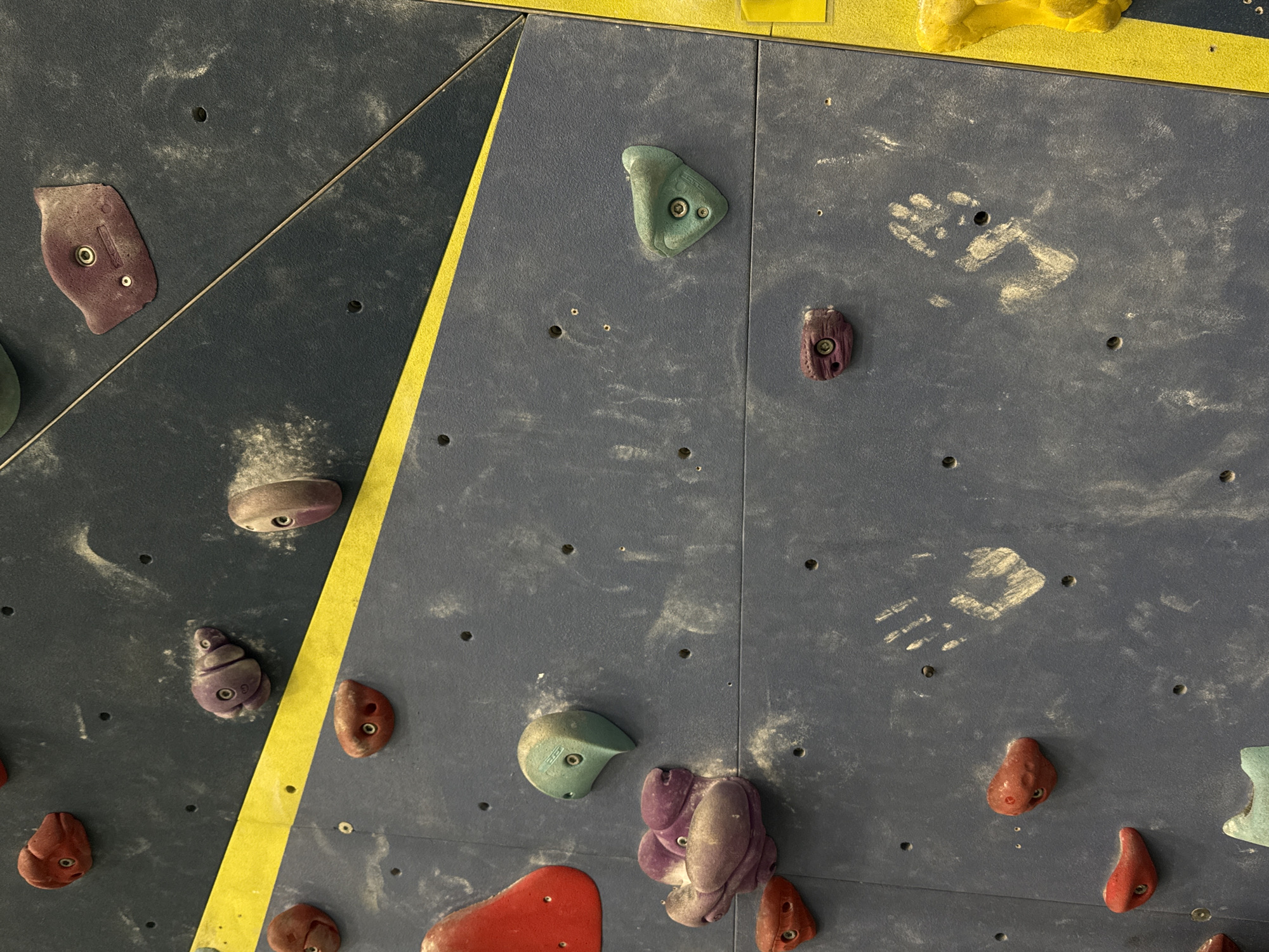 The image shows a section of an indoor rock climbing wall. The wall is constructed from blue panels, some bordered by yellow trim. Various climbing holds in different shapes, sizes, and colors (red, purple, green, and yellow) are bolted onto the wall. Chalk smudges and handprints are visible on the blue surface, indicating use. Based on the climbing holds and overall appearance, this is likely a facility within Sofia, Bulgaria, as that location was specified. No people are visible in the frame.
