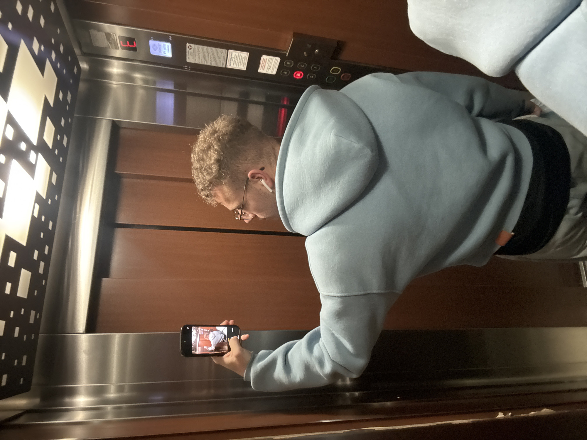 The image captures a young adult male inside an elevator, rotated 90 degrees counter-clockwise from a standard vertical orientation. The man has light brown, curly hair, wears glasses, and has a white earbud in his left ear. He is dressed in a light blue hooded sweatshirt and dark pants.
He is actively engaged with a black smartphone, holding it horizontally in his right hand, pointing it towards himself. The phone's screen displays a live preview, seemingly a selfie or video of himself, showing his reflection in the light blue hoodie.
The elevator's interior features warm brown wooden panels on the rear and right walls, contrasted with shiny brushed stainless steel panels, particularly noticeable at the base and between wall sections. The left wall features a dark panel with a geometric pattern of illuminated white squares and rectangles, likely a decorative light fixture. Part of light-colored, upholstered seating or cushions is visible in the upper right portion of the frame.
Above the wooden panel on the right is the elevator's control panel. A digital display shows "E3", indicating the elevator is on the third floor or an equivalent level. Below this, numerous floor buttons are visible, with the "2" button prominently lit in red. Other buttons include a red bell icon and controls for opening and closing doors. Several warning or informational placards are affixed to the panel; while most text is illegible, one appears to have a diamond-shaped warning symbol. The setting is clearly indoors, illuminated by artificial light. No specific location cues are visible within the image to confirm Plovdiv, Bulgaria. The man appears focused on his phone, with no discernible emotional expression.
