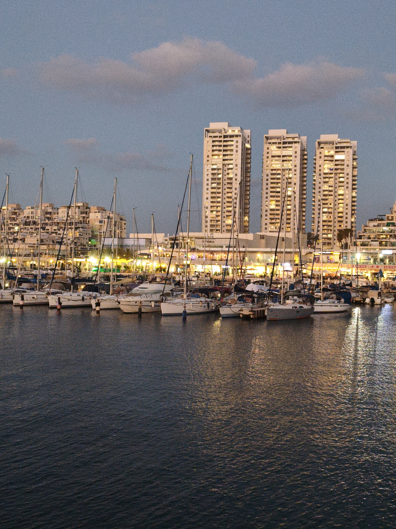 The image depicts the Ashkelon Marina in Ashkelon, Israel, at dusk. The scene features a body of water with numerous sailboats docked. Behind the boats, a complex of modern, tall buildings with illuminated windows lines the waterfront, suggesting a residential or commercial area. The sky above is a soft blue with scattered clouds. The lighting indicates that it is transitioning from daylight to evening, with artificial lights from the buildings and boats beginning to glow. The water's surface reflects the lights, creating a shimmering effect. No people are clearly visible in the image.
