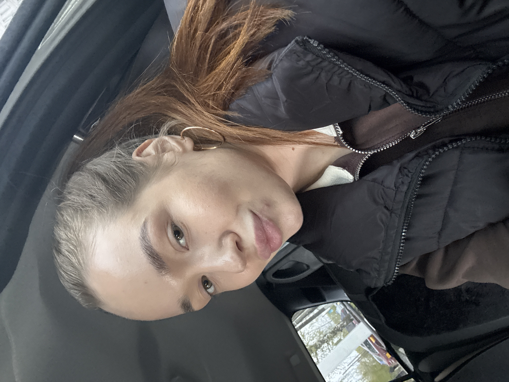 A woman with auburn hair and fair skin is seated inside a car. She is wearing a black puffer jacket over a brown top. The steering wheel of the car is visible in the background, as well as the car window. Outside the window, there are signs of a city location, possibly Chaikovsky, Russia, though there are no immediately clear landmarks to confirm the location.