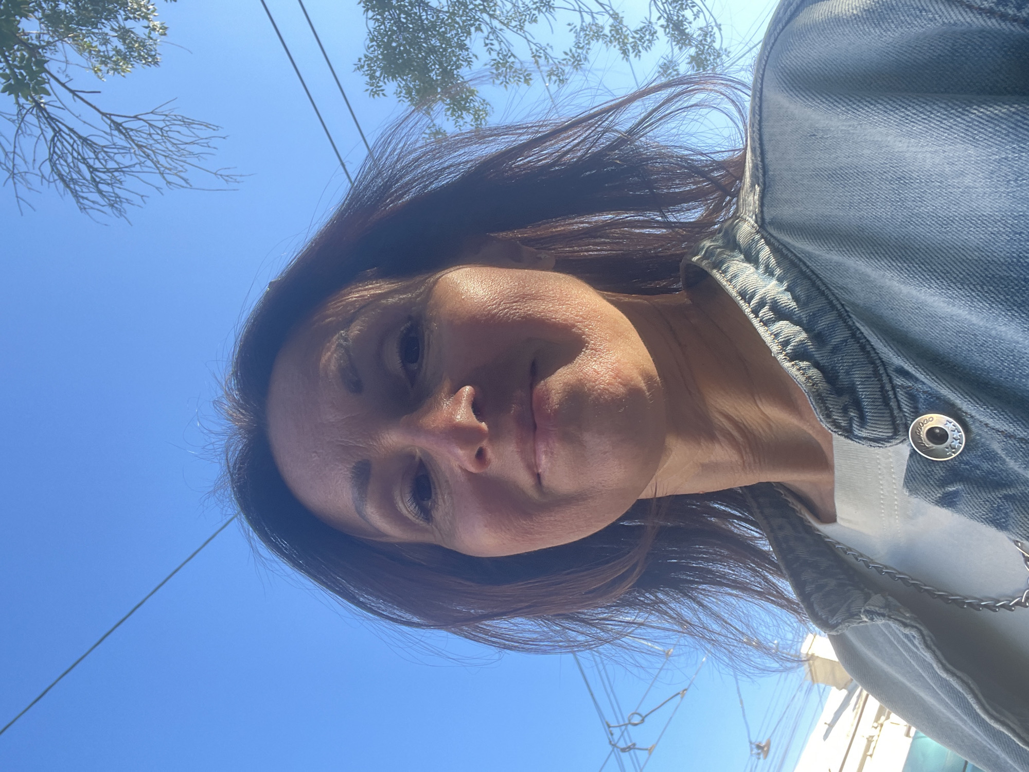 A medium shot of a woman facing the camera, her head tilted slightly back. She has long, dark brown hair, fair skin, and appears to be outdoors under a clear blue sky. The woman is wearing a light-colored t-shirt and a denim jacket. A silver chain necklace is visible over the t-shirt. The background shows some tree branches and what appear to be electrical wires, suggesting an urban environment. There is no visible indication of the city being Saint Petersburg, Russia.