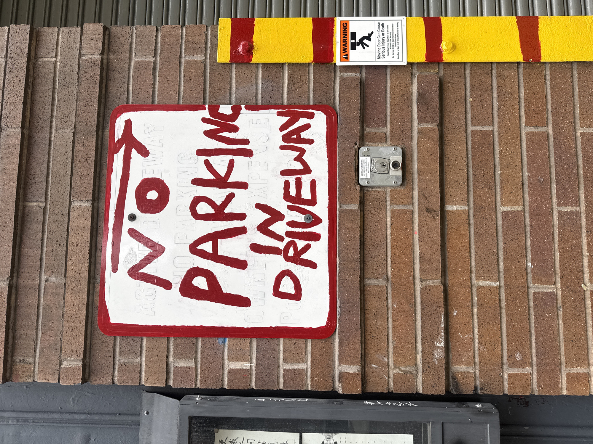 The image depicts a section of a brick wall with signage. A "No Parking Driveway" sign is prominently displayed with a red border and red lettering, indicating a restricted parking area. Above the sign, there's a yellow bar with red stripes, possibly a safety barrier or marker. Affixed to this bar is a warning label containing an icon of a person being crushed, cautioning about the dangers of a moving door and potential serious injury or death. A small security device, possibly a keypad or entry system, is mounted between the sign and the yellow bar. The location is indicated by the text "M&M GATE SERVICE" and the phone number "917-757-2420", which suggests the scene is located in New York City, United States of America, due to the 917 area code. The presence of the gate service company also indicates the location might be near a gated area or entryway.
