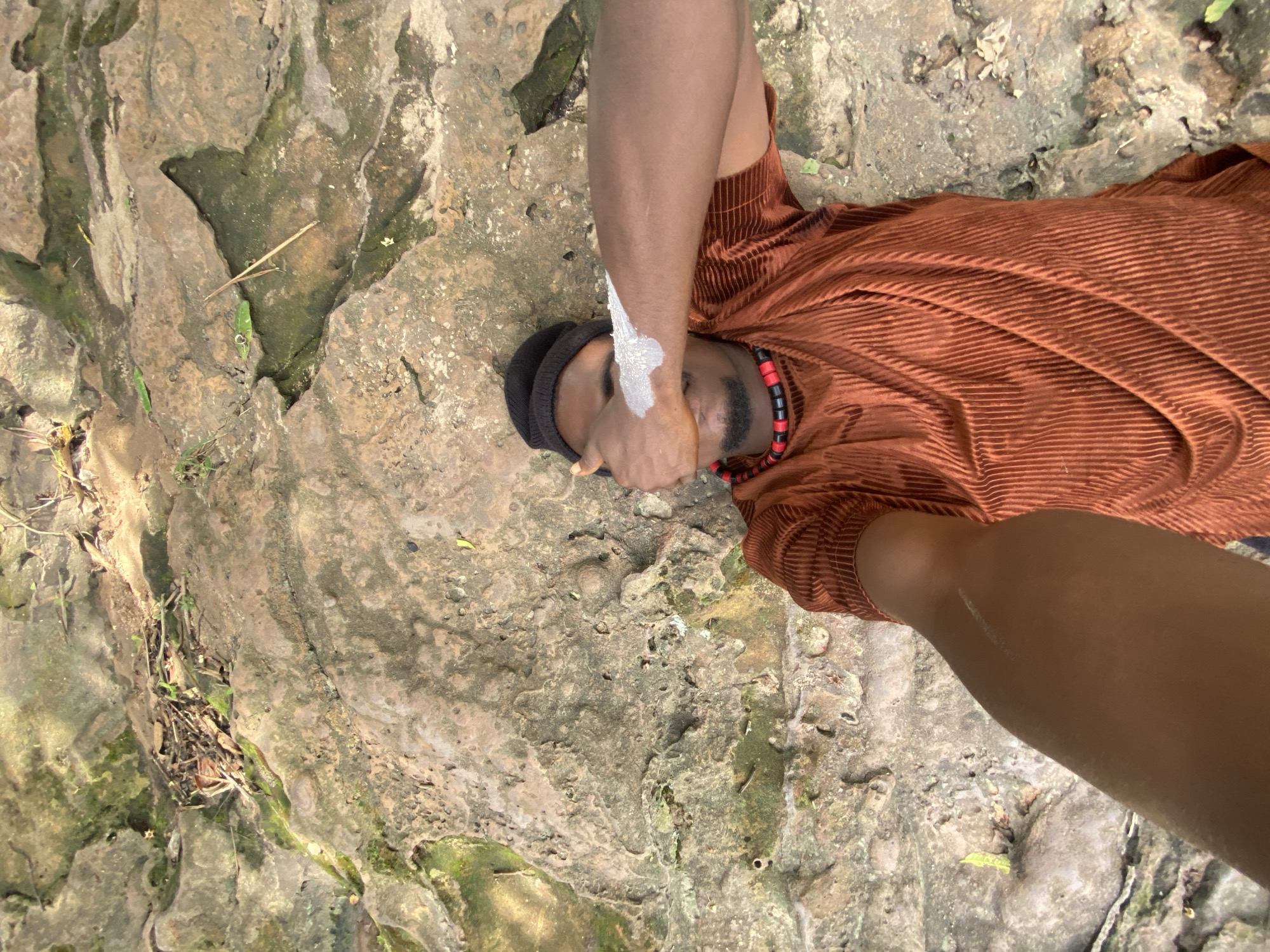 The image shows a person lying on a mossy, rocky surface. The person is wearing a rust-colored, horizontally striped shirt and a black beanie. They have a red and black beaded necklace. White substance is smeared on their arm and forehead. The scene is outdoors, likely in a natural area, as the rocky surface suggests a natural environment. The location may be near Igbara, Nigeria, based on the context provided.
