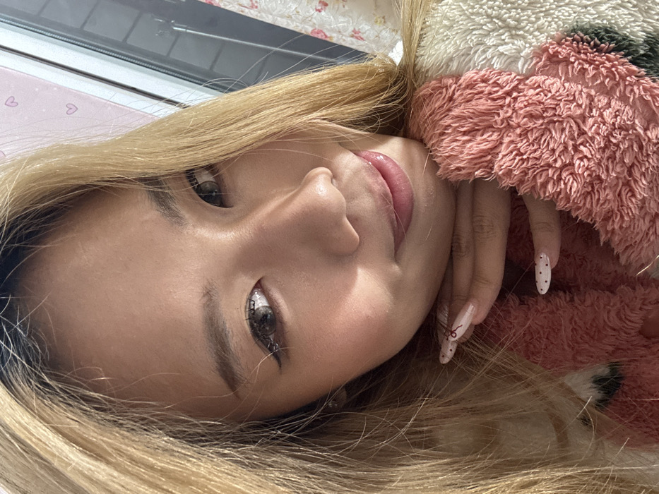 The scene is an indoor setting within Shapingba District, China. A close-up portrait features an East Asian female. She has long, blonde hair with dark roots and grey-brown irises, consistent with contact lens use. Visible makeup includes eyeliner and blush. Her right hand, displaying elongated, light-colored artificial nails—some with dark polka dots and one with a red linear design—is positioned beneath her chin. She is wearing a fluffy garment with visible pink and white sections. Lighting from the right illuminates the subject's right side, casting a shadow across her left cheek. A reflective surface, possibly a window, is partially visible on the right, revealing a pink background adorned with small heart motifs. The subject looks directly forward with a slight smile.