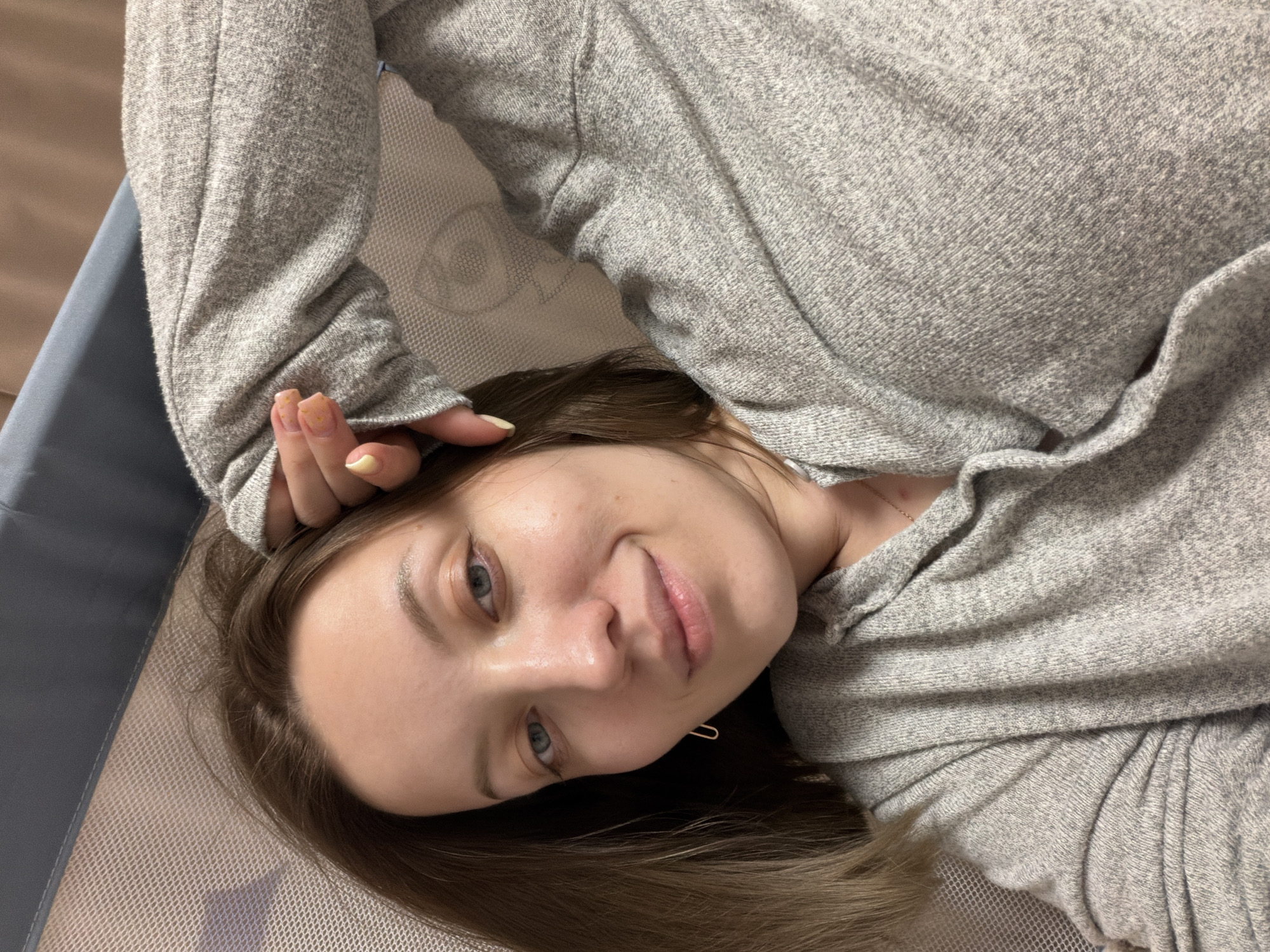 A young woman is shown lying down, captured from a slightly elevated perspective, in an indoor setting in Reshetnikovo, Russia. She possesses fair skin, light blue eyes, and long, flowing medium-brown hair that frames her head. Her expression is serene and relaxed, featuring a gentle, subtle smile.
She is dressed in a light grey, long-sleeved top, characterized by a soft, heathered or subtly flecked knit fabric. A delicate gold chain with a small, minimalist pendant is visible around her neck. Her left hand is positioned near her head, revealing manicured nails painted in a light yellow-gold hue with tiny, decorative glitter or star details.
The woman rests on a light-colored, possibly cream or white, mesh or finely woven surface, suggesting a bed, mattress, or perhaps a baby playpen. To her left, a structural element with a dark blue or grey fabric border is visible, reinforcing the impression of a designated resting area. The background is softly blurred, indicating an indoor domestic environment. The lighting is soft and diffused, consistent with interior daytime conditions. No text is visible in the image.
