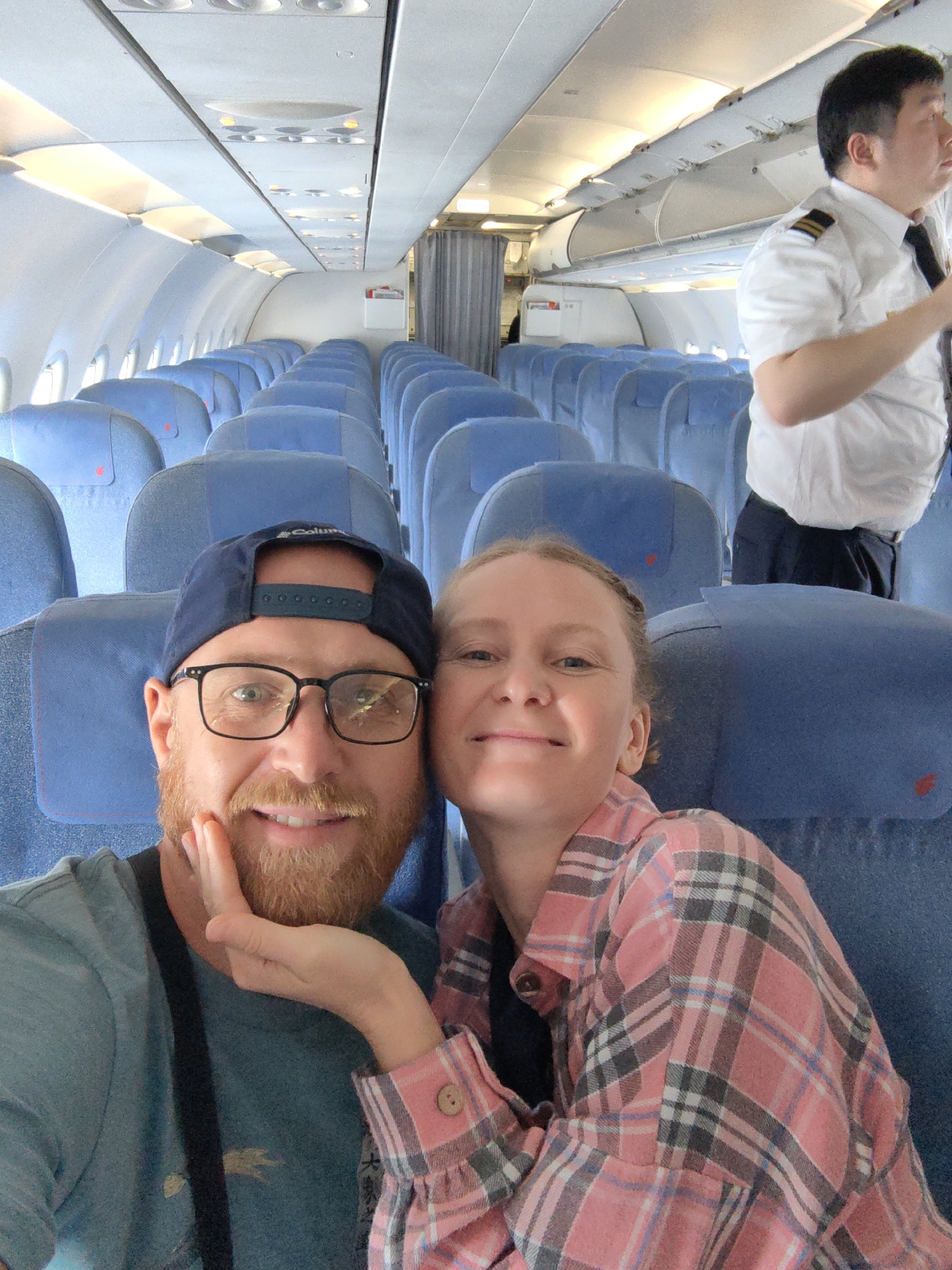 A selfie taken inside an airplane shows a man and a woman sitting in blue seats. The man is wearing glasses and a blue baseball cap. The woman is smiling and wearing a pink plaid shirt. In the background, the empty seats of the airplane are visible, and a man in a pilot uniform is standing in the aisle. The image does not provide information about the city of Hà Nội, Vietnam.