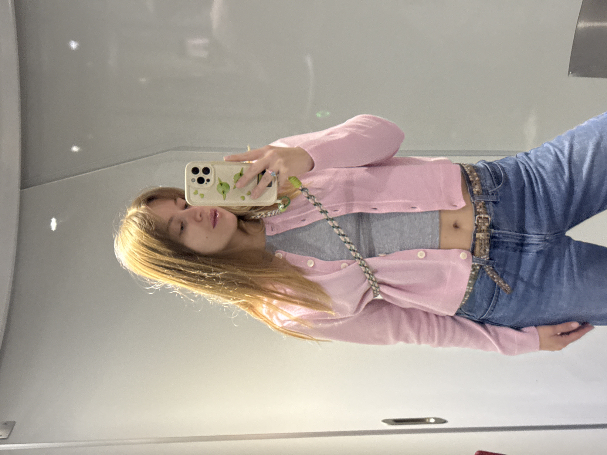 A woman is taking a mirror selfie in what appears to be a public restroom, based on the style of the mirror and other visible fixtures. She is wearing light blue jeans with a brown belt, a gray crop top, and a pink cardigan. She holds a phone with a cream-colored case featuring a design of small green fruits or leaves. The location is Unknown, Unknown.