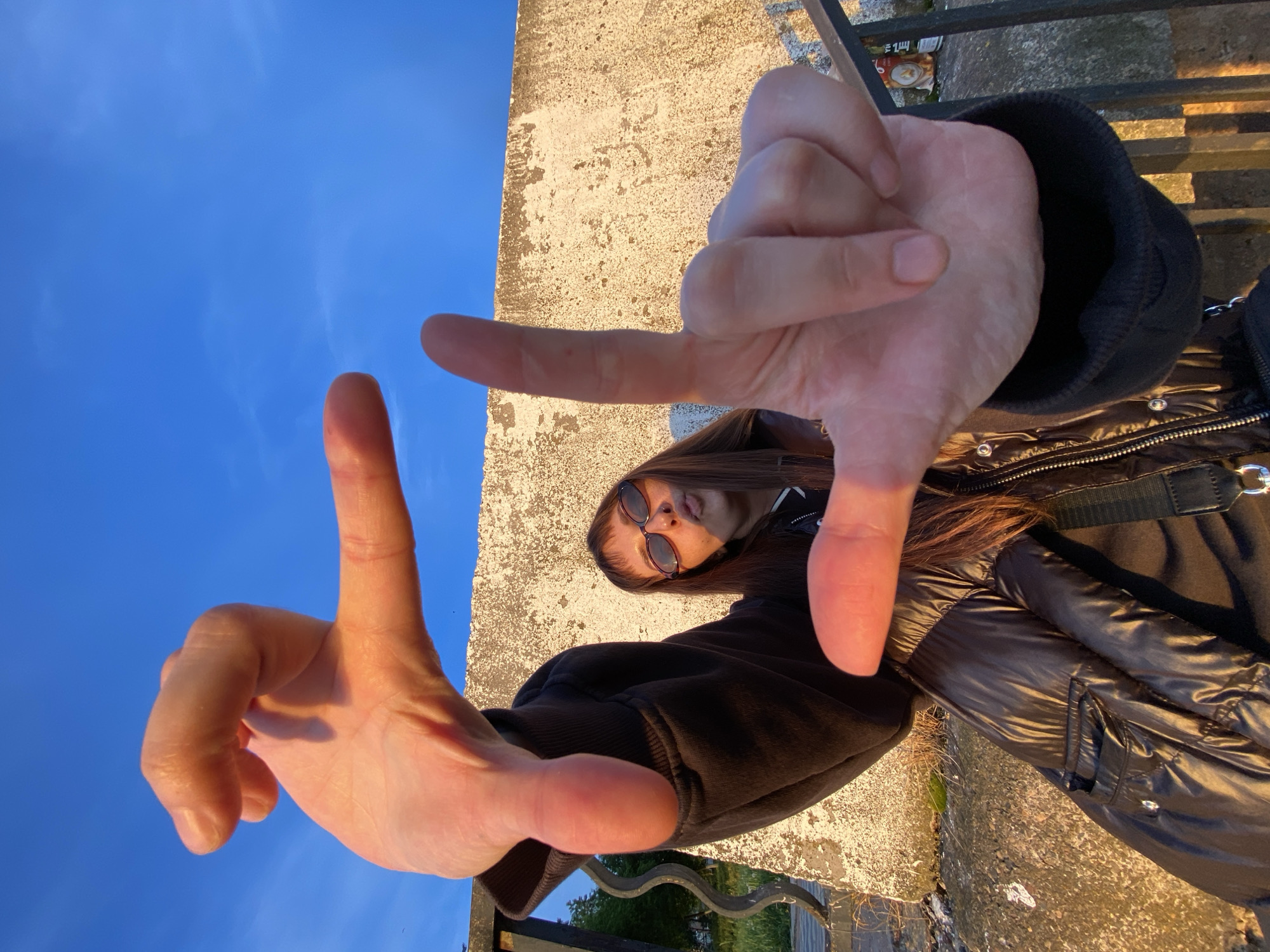 A young woman with long brown hair and sunglasses is centered in an upward-facing shot, framing her face with her hands in a loose rectangle. She is wearing a black puffer jacket and a black hooded sweatshirt. The background includes a blue sky, a concrete wall with a rough texture, and a black metal railing. Based on the architectural details, this image was likely taken in Votkinsk, Russia.