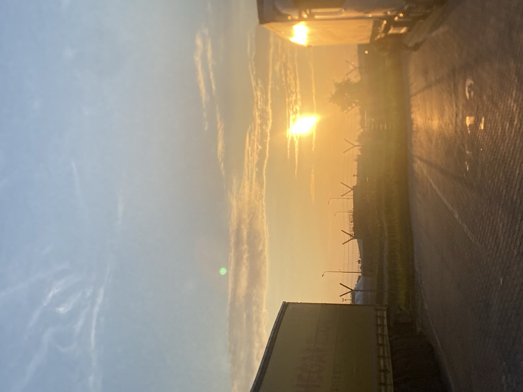 The image depicts a sunset at what appears to be a logistics or industrial area in Niepołomice, Poland. The sky is a gradient of pale blue and orange, with wispy clouds. The sun, a bright, elongated orb, is low on the horizon, casting a warm glow and creating reflections on the wet pavement in the foreground. Two large trucks are visible, one yellow on the left and a white one on the right, parked side-by-side. Their trailers are partially cut off by the frame. In the background, there are industrial structures, including fences with Y-shaped posts, streetlights, and possibly buildings. The ground is paved with interlocking bricks, and there are puddles of water. No people are visible in the scene.