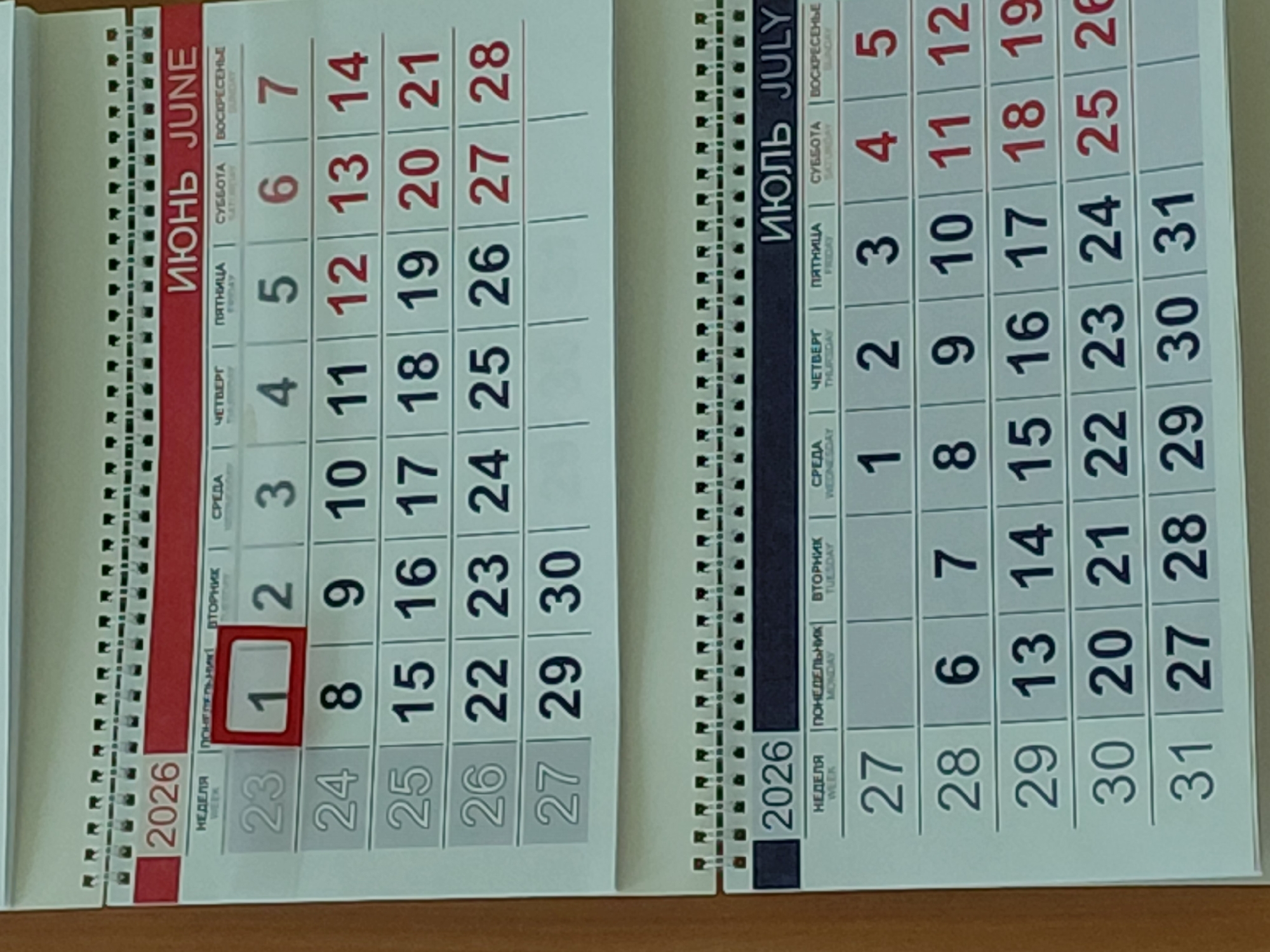 The image shows a desk calendar displaying the months of June and July 2026. The June page is on the left, with the number "1" highlighted in red, indicating the start of the month on a Tuesday. The July page is on the right, showing the beginning of the month on a Wednesday. The calendar is written in both Russian and English. The year "2026" is printed on both pages. The calendar appears to be on a wooden surface.