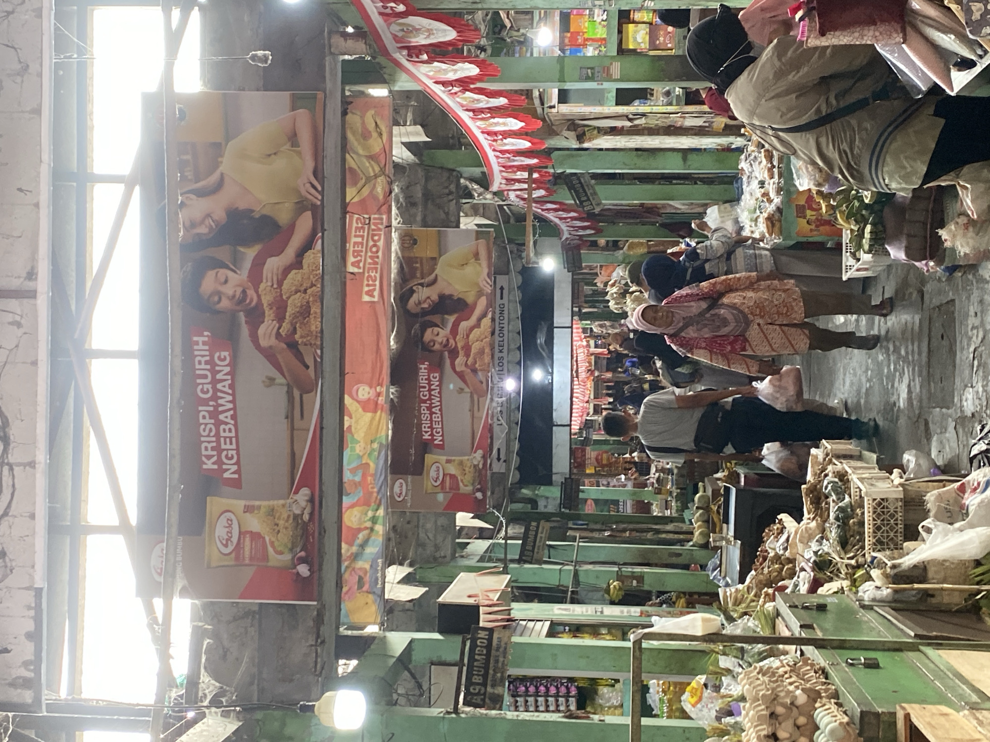 The image shows a bustling marketplace in Ambarketawang, Indonesia. In the foreground, a person wearing a headscarf and a patterned jacket walks towards the viewer. To their left, various goods, including eggs and produce, are displayed on stalls. In the midground, people are seen browsing stalls laden with food items, some covered with plastic. A sign for "LOS KELONTONG" is visible, indicating a general store. The background reveals a long corridor of market stalls, with the ceiling and upper structures of the market visible. Two large banners advertising a food product with images of people eating are prominently displayed overhead. Red and white pennants are strung across the upper part of the market. The overall scene depicts a vibrant and active marketplace environment.