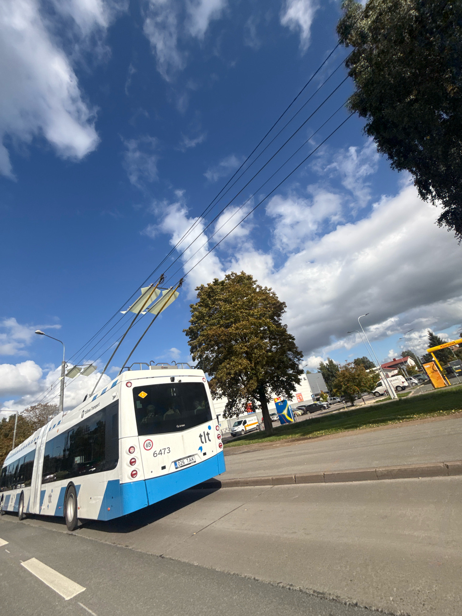 The image shows the rear and side of a white and blue trolleybus with the markings "Tallinn Transport" and "tlt" on its rear. The trolleybus has the number 6473 and license plate "026 TAK" visible. It is on a street in Tallinn, Estonia, with a sidewalk and a grassy area with trees and buildings in the background. The sky is blue with white clouds. Overhead power lines for the trolleybus are visible. A traffic speed limit sign of "65" is also present. The scene appears to be daytime, with sunlight casting shadows.