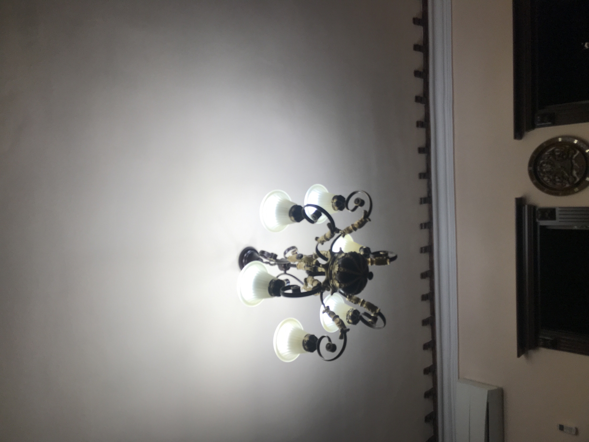 The image shows a ceiling with a chandelier in a room. The chandelier is ornate, with multiple lights arranged in a circular pattern. To the right, there is a wall with dark-framed objects and a decorative molding. The location is unknown; there is no information to determine if the photo was taken in Yerevan, Armenia.