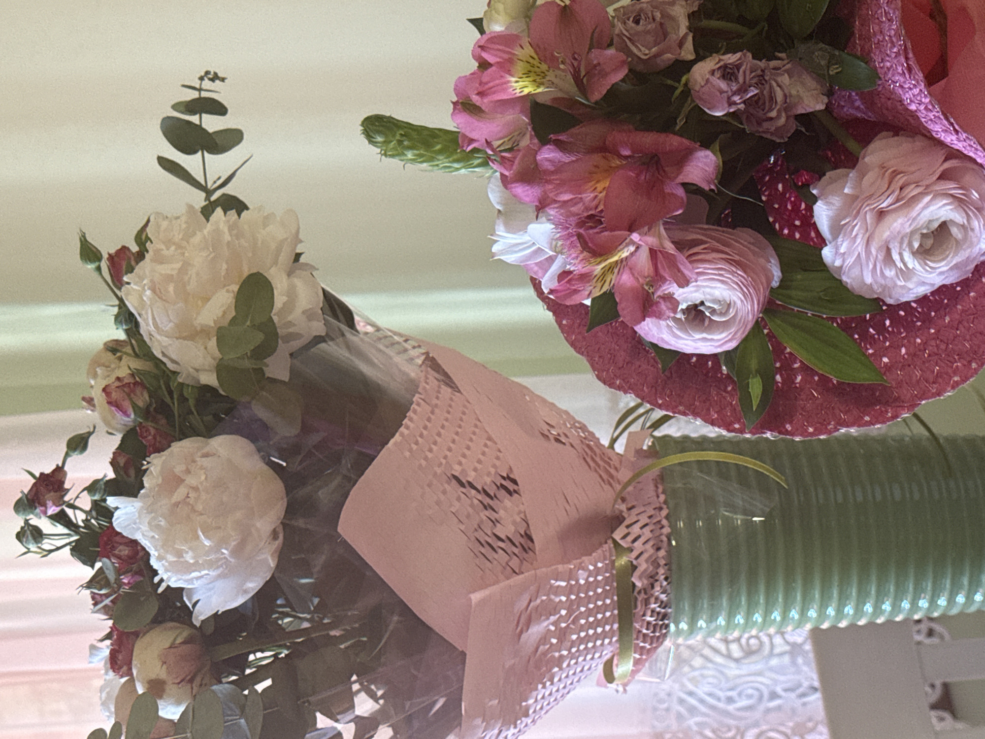 Two bouquets of flowers are arranged in vases. One bouquet features white peonies and small red roses wrapped in pink paper. The other bouquet is composed of pink alstroemeria, roses, and ranunculus in a pink vase. The vases sit on a white table with a patterned surface. The background is a plain, light-colored wall. The location is not specified and is not possible to determine based on the visual information alone.