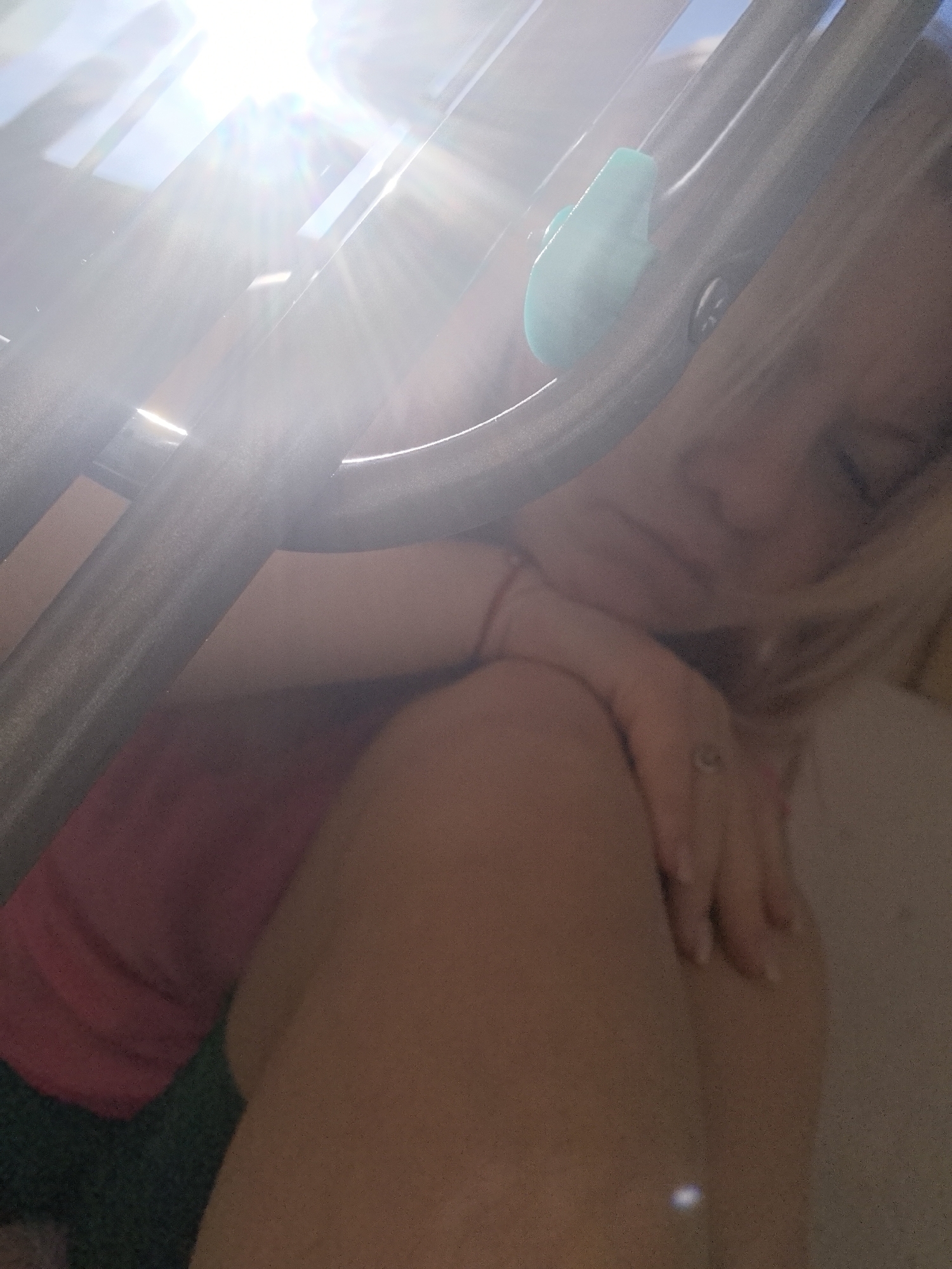 The image shows a close-up view of a woman's arm and shoulder, draped over a metal railing. The sun is shining brightly, creating lens flare and overexposure in the upper part of the image. The woman appears to be wearing a pink top and a bracelet on her left wrist. A small turquoise object is attached to the railing near her arm. The background is blurred, but hints of a blue sky and green foliage suggest an outdoor setting. The location is identified as Chaikovsky, Russia.