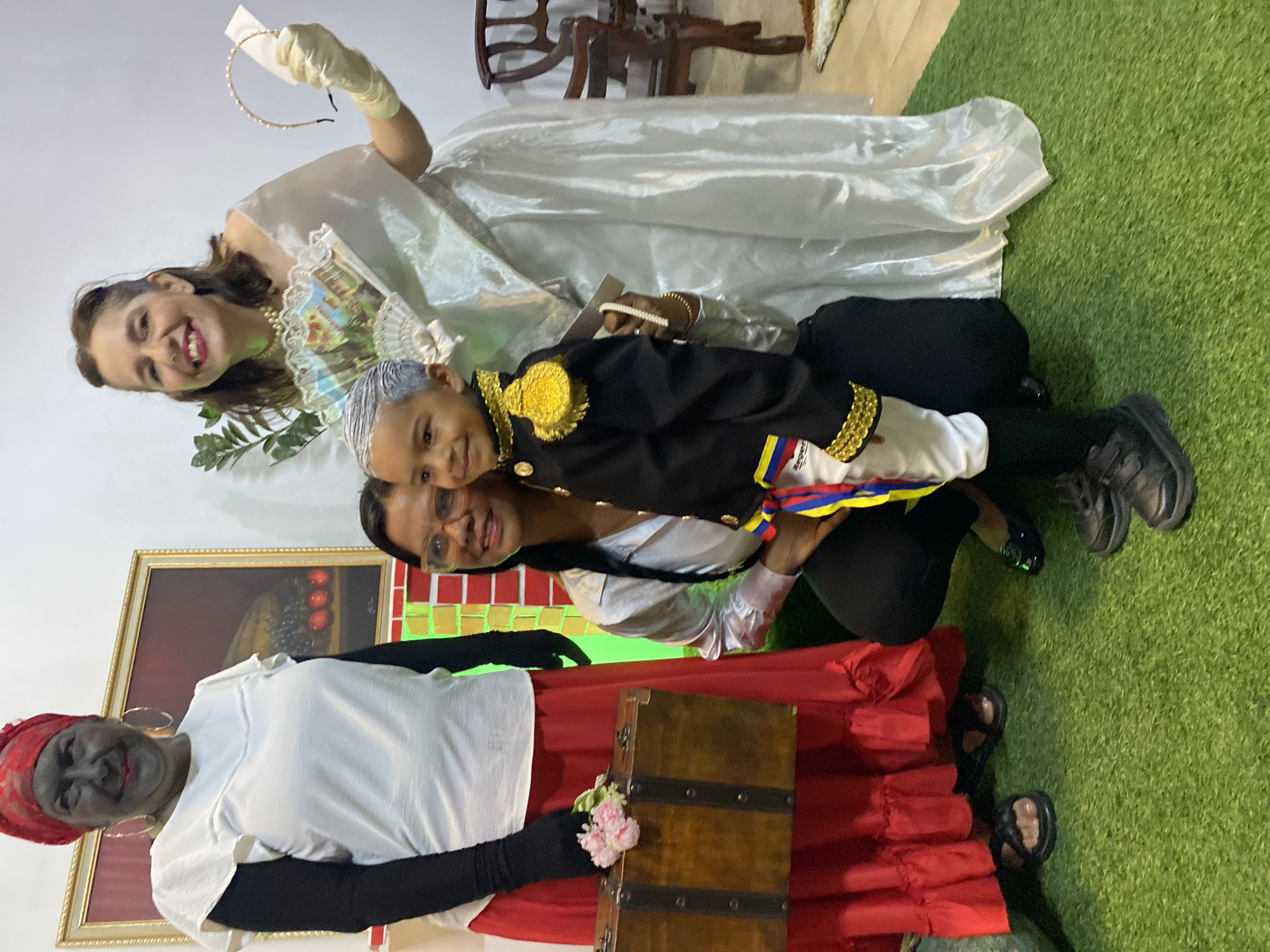This image captures four individuals—three adults and one child—posing indoors in Tinaquillo, Venezuela. The setting features an artificial green turf floor and white walls.
On the left, an adult woman stands, her face covered in dark makeup, adorned with a red headwrap, a white blouse over a black long-sleeved undershirt, and a ruffled red skirt. She wears black sandals and holds a small, rectangular wooden trunk with metal clasps, topped with a small cluster of pink flowers. Behind her, a framed painting with dark tones and a depiction of red objects is visible, along with a structure that appears to be illuminated with green and red lights.
In the center, another adult woman, wearing eyeglasses, a silver-colored collared shirt, and black pants, crouches. Her left arm is around a young boy positioned in front of her. The boy, who has his hair painted grey, is dressed in a black military-style jacket with gold trim and epaulets, white pants, and a ribbon in the colors of the Venezuelan flag tied around his waist. He wears black sneakers.
To the right, a third adult woman stands, smiling. She wears a white, off-the-shoulder gown, white gloves, and a pearl necklace. She holds a decorative fan with a landscape image and a pearl-beaded headband in her gloved left hand. A green leafy plant and a partial wooden chair are visible in the background behind her. All four individuals are smiling and looking towards the camera.