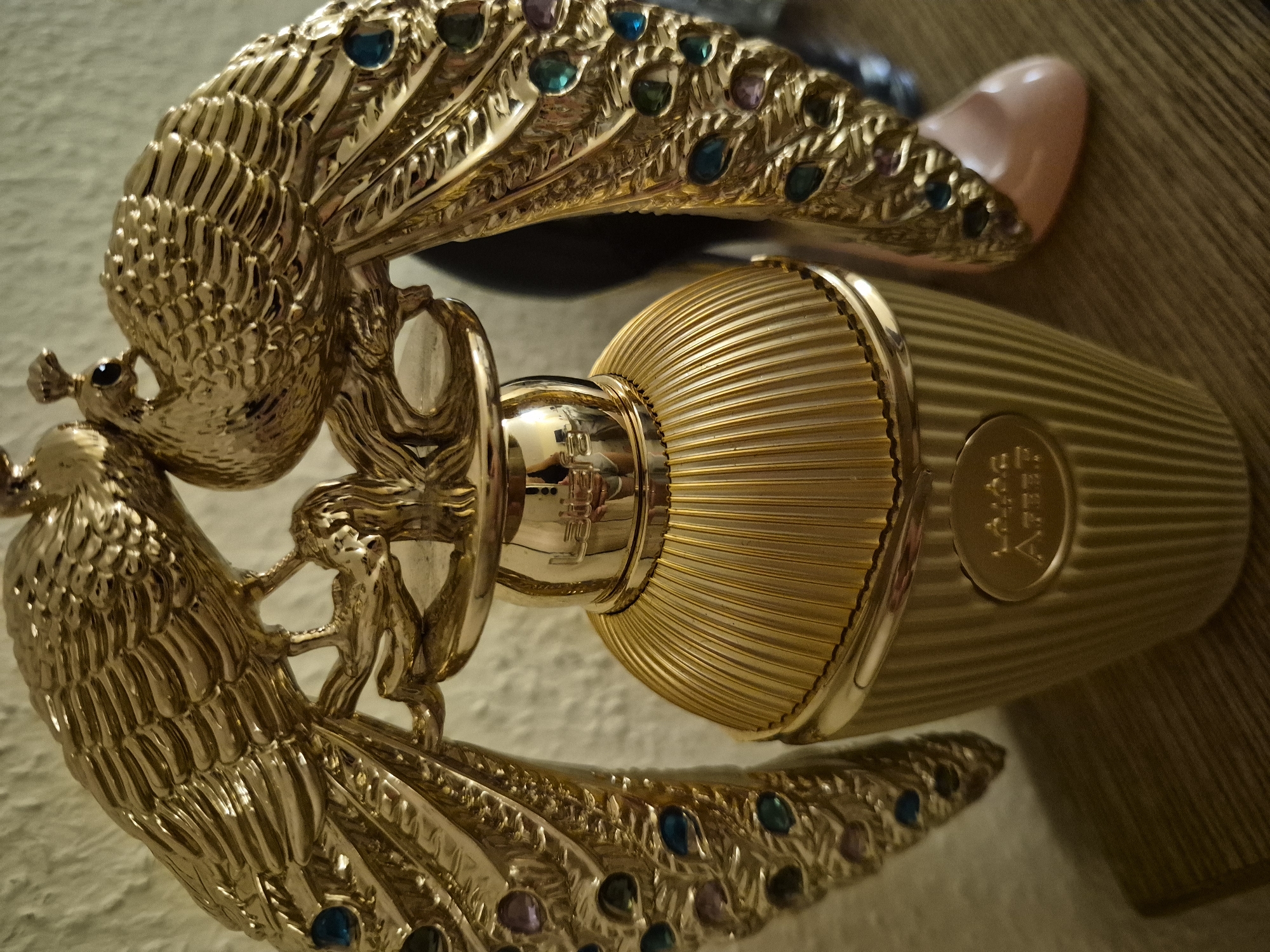 A gold-colored perfume bottle with a fluted design stands on a wooden surface. The bottle is topped with a decorative gold sculpture of two peacocks facing each other, with their tails fanned out and adorned with colored jewel-like accents. The bottle itself has a circular emblem near its base with some writing on it. The background is a textured wall. The scene is lit from above, casting shadows on the surface and wall.