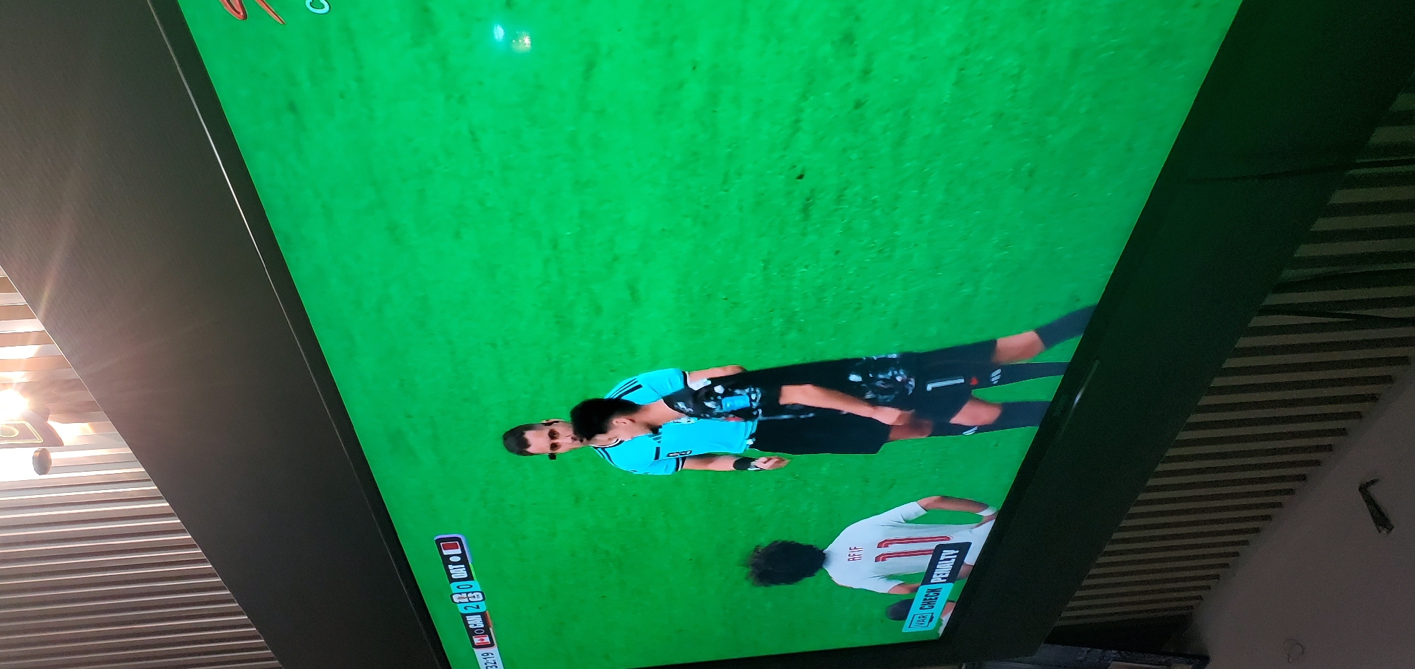 This is a photograph of a television screen displaying a soccer (football) match. The screen shows a green grass field, indicating an outdoor sports venue. In the foreground, two men are visible on the field. One man, wearing a light blue jersey with black accents and shorts, appears to be a referee due to his attire and proximity to another player. He has dark hair and is looking down. The second man, also with dark hair, is wearing a black jersey with white numbering and black shorts. He seems to be engaged in a conversation or interaction with the referee.
Further in the background, another player is visible wearing a white jersey with the number 11 prominently displayed and the name "RIFF" above it. This player also has dark, curly hair.
At the bottom left of the screen, a score overlay is partially visible. It shows "32:19" which likely indicates the current time in the match. To the left of this time, there are some flags and team identifiers, but they are not entirely clear. One flag appears to be for Canada, indicated by a red maple leaf. Another flag, possibly for Saudi Arabia, is also visible. The score is displayed as "2" next to the Canadian identifier and "0" next to the other team's identifier.
On the right side of the screen, a "VAR CHECK PENALTY" notification is displayed, suggesting a crucial moment in the game where the Video Assistant Referee is being consulted. The overall scene conveys a moment of intense focus and potential decision-making during a soccer match. The lighting appears to be from artificial sources, typical for indoor viewing or an overcast day.