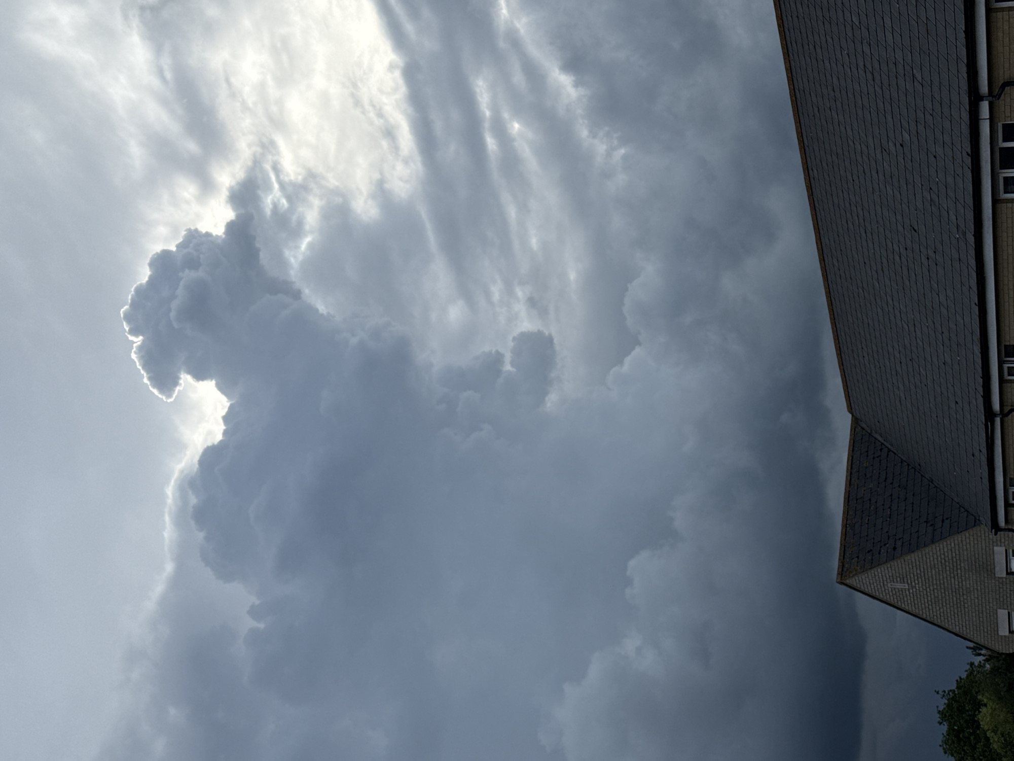 The vertically oriented image, when viewed upright, presents a dramatic and dynamic sky with a building partially visible on the right. The upper left and central portions of the sky are dominated by brilliant, luminous clouds, strongly backlit by an unseen light source, likely the sun, creating bright white edges and a glowing effect. These clouds are dense and textured, appearing powerful and almost three-dimensional. Towards the center and right, the sky darkens significantly, filled with heavy, turbulent, dark grey and blue-grey clouds, indicative of stormy or rapidly changing weather conditions.
On the right side, the dark, steeply sloped roof of a building cuts diagonally across the frame. The roof is covered with numerous small, dark, rectangular tiles or slates, creating a patterned surface. Below the roofline, a section of a light-colored building wall, possibly brick or rendered, is visible, featuring a few small windows with white frames and a vertical downspout. A small cluster of dark green foliage is visible at the very bottom right, partially obscured by the dark clouds and building. The scene captures a moment of intense atmospheric activity, likely during the day, with the contrasting light and dark clouds suggesting an impending or recent storm over an urban or suburban area, consistent with the Great Yarmouth, UK context. No people or text are visible.