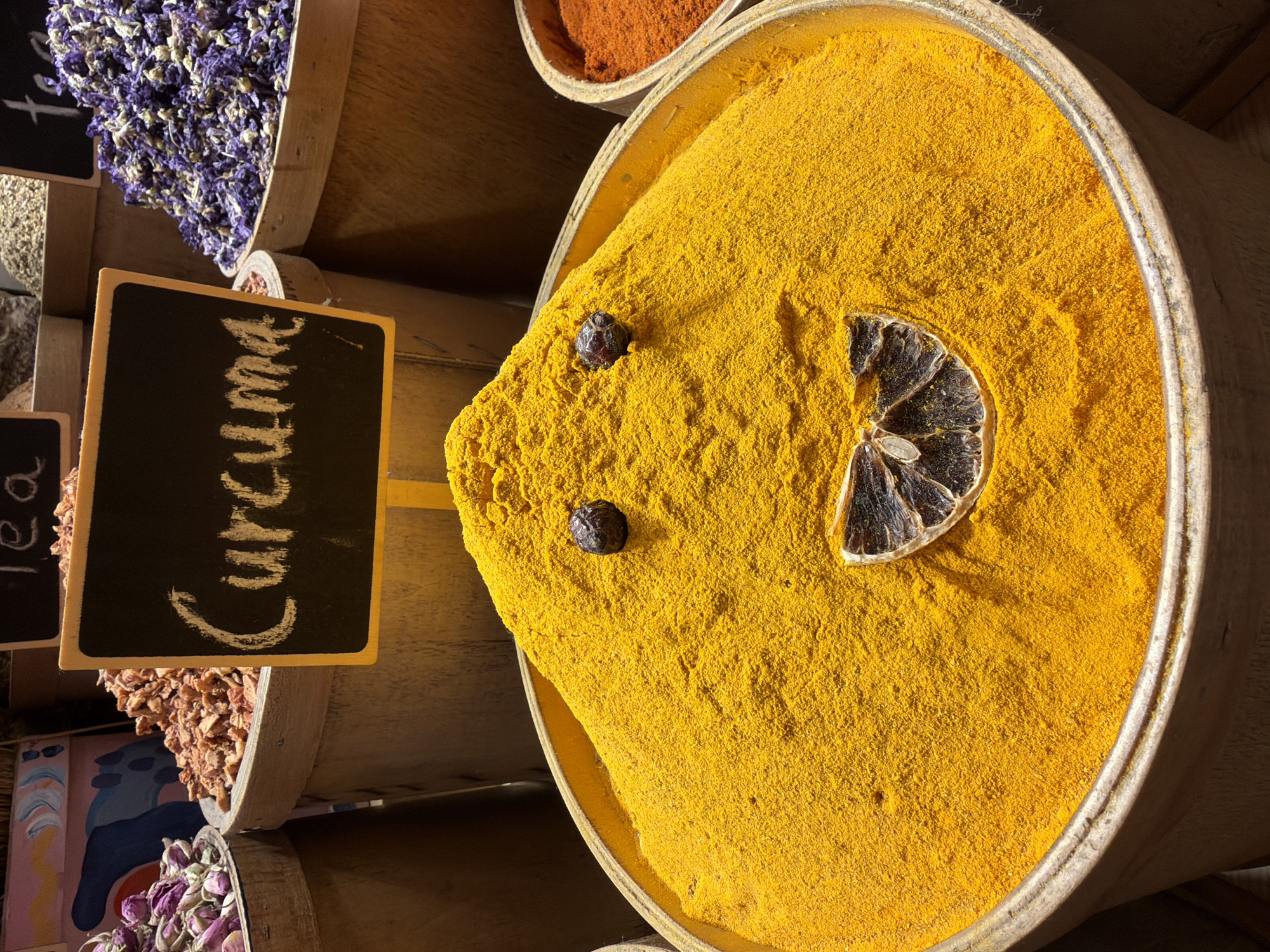 This media file presents a close-up, eye-level view of barrels filled with spices, likely in a market setting such as Istanbul, Turkey. The primary focus is on a large barrel brimming with vibrant yellow turmeric powder, forming a mound with a slightly sunken appearance. Embedded in the turmeric are two small, dark, whole spices, possibly peppercorns or cloves, and a dried slice of citrus fruit, positioned to resemble a decorative element.
To the left of the turmeric barrel, another barrel is visible, containing a heap of dried purple flowers, likely lavender, suggesting a variety of spices and botanicals are being sold. A small, black chalkboard sign with a wooden frame is propped against the flower barrel, displaying the word "Curcuma" in white chalk handwriting. Below this sign, another barrel holds a dried, reddish-brown granular spice. Further down, a glimpse of a barrel with dried rosebuds is visible, adding to the rich display of natural products.
The lighting suggests an indoor setting or a shaded outdoor area, casting subtle shadows that highlight the textures of the spices. The overall impression is one of a traditional spice bazaar, abundant with color and natural ingredients. The visible text confirms the presence of turmeric and hints at other potential offerings. There are no people visible in the frame, and the scene appears to be static, focused solely on the arrangement and presentation of the spices.