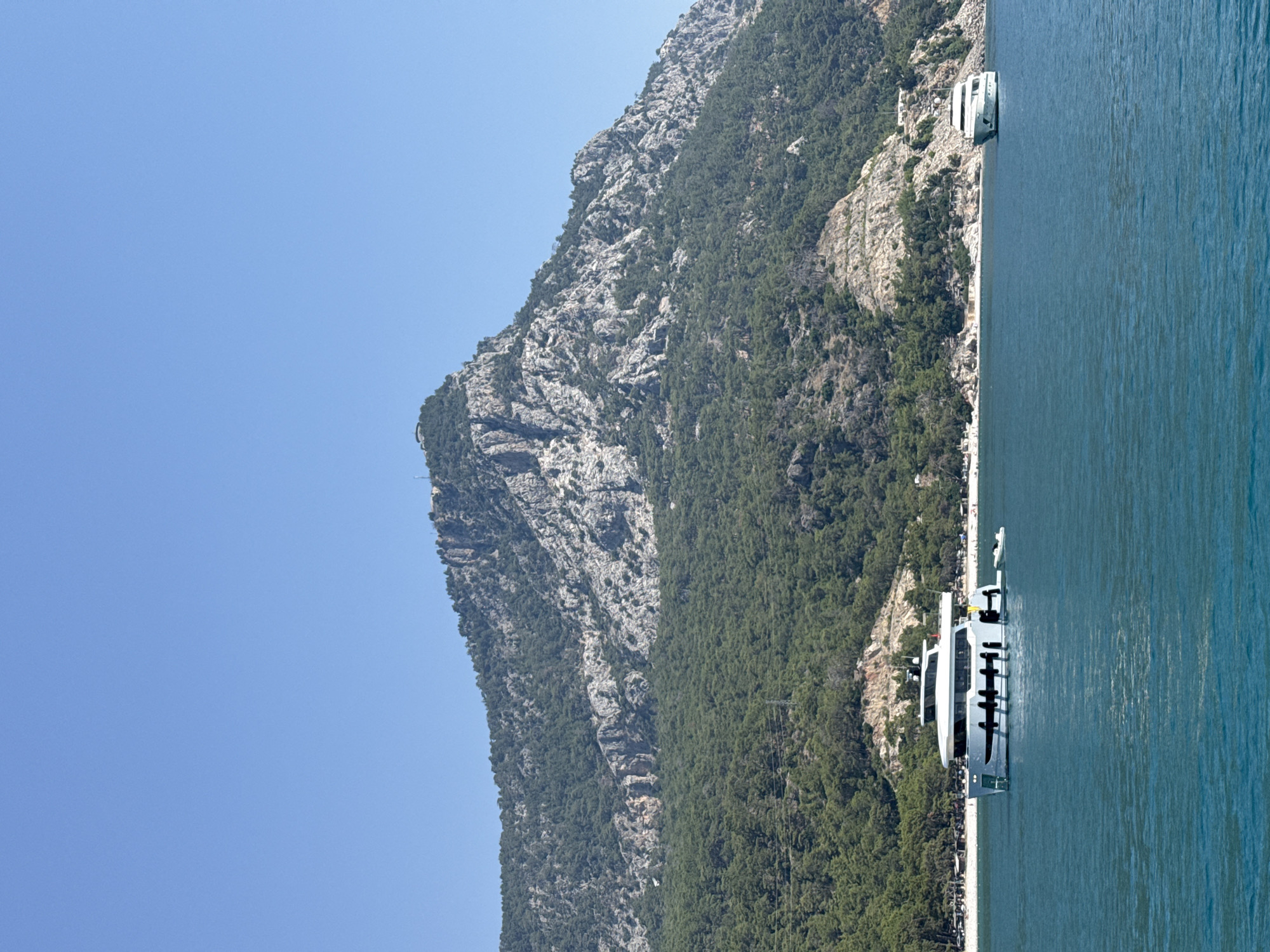 The image depicts a panoramic view of a coastal mountain range meeting a body of water. The mountain is covered in dense green foliage, with rocky outcrops and sheer cliffs visible. A clear blue sky stretches across the upper portion of the frame. Two white boats are moored near the shore. The boat in the foreground is a modern yacht with multiple decks and windows. The second boat, further in the distance, appears to be a smaller vessel. The water is a deep blue, with subtle ripples on the surface. A narrow strip of land, possibly a beach or walkway, separates the mountain from the water. The scene suggests a sunny day with calm weather, ideal for boating or observing the natural landscape. The overall impression is one of natural beauty and tranquil leisure. No visible text is present in the image.